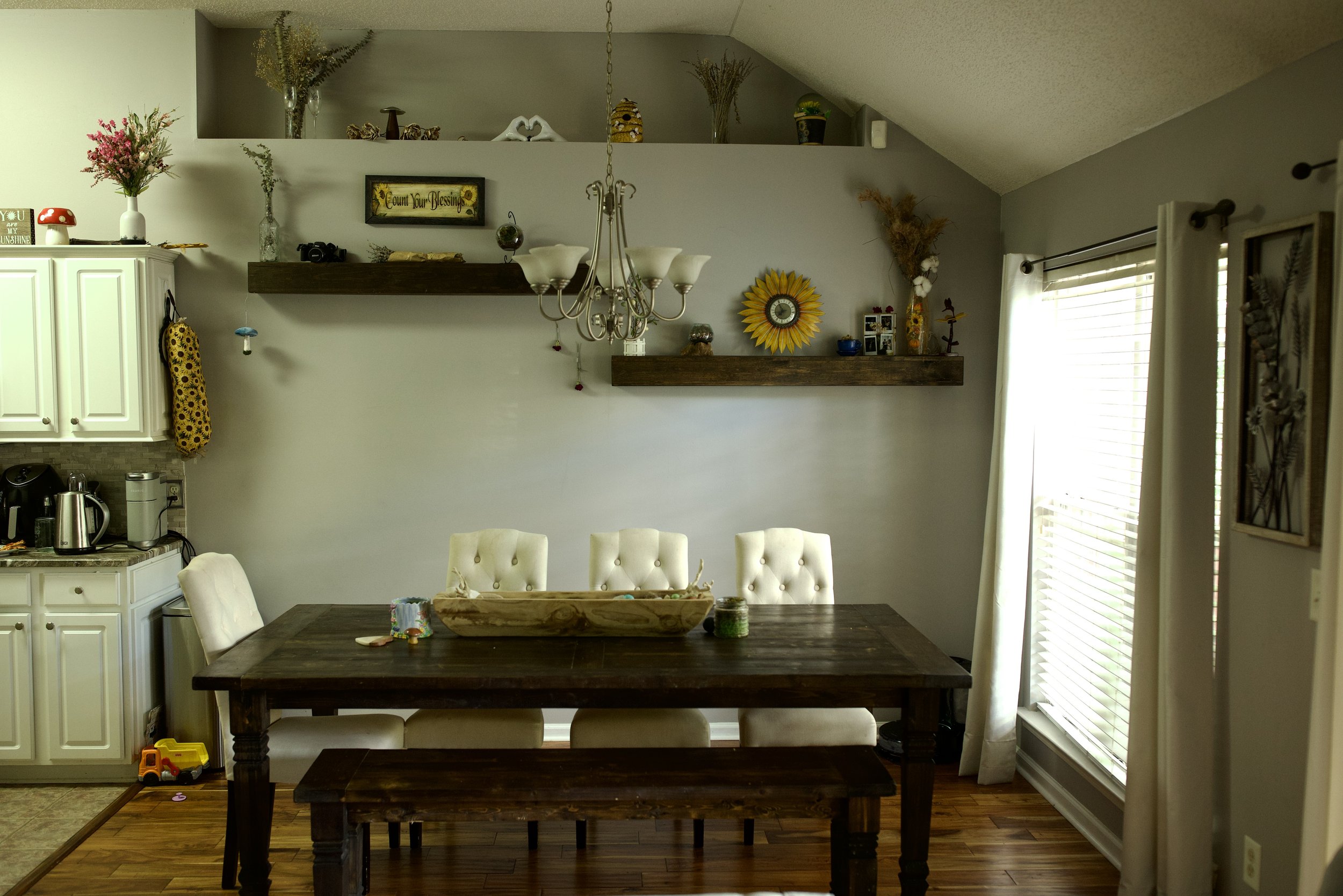 A dining room with a wooden table and six white upholstered chairs. There are two wooden shelves with decorative items, including vases, a sunflower wall clock, framed photos, and plants. A chandelier hangs above the table. To the right, there are la