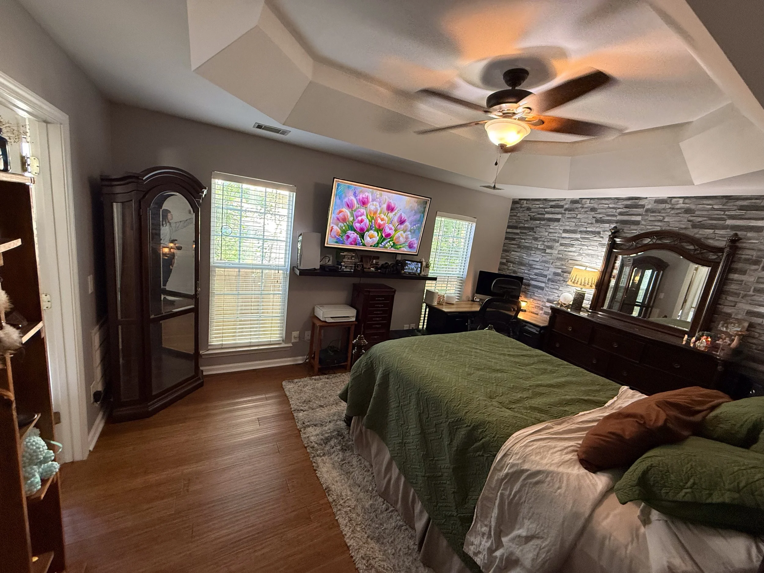 A bedroom with two large windows, a stone accent wall, a ceiling fan, a large mirror, a TV, and a bed with a green quilt and brown pillows.