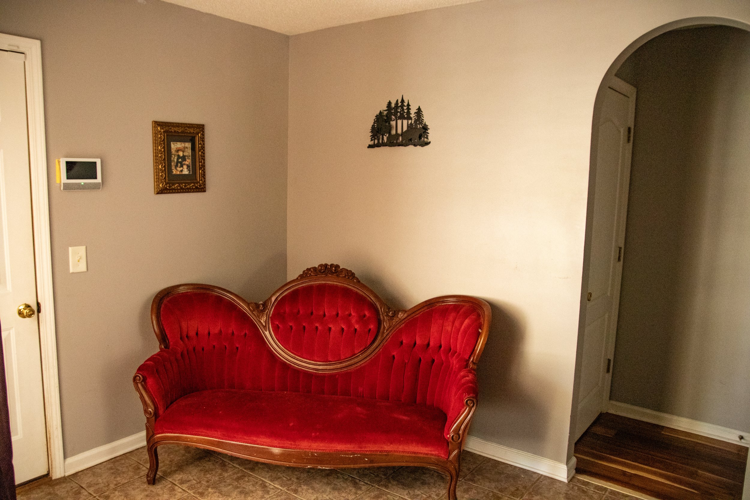 Interior of a house with a vintage red velvet sofa, wall decorations including a framed picture and a small wall sculpture of trees, a thermostat, a door, and part of an archway leading to another room.