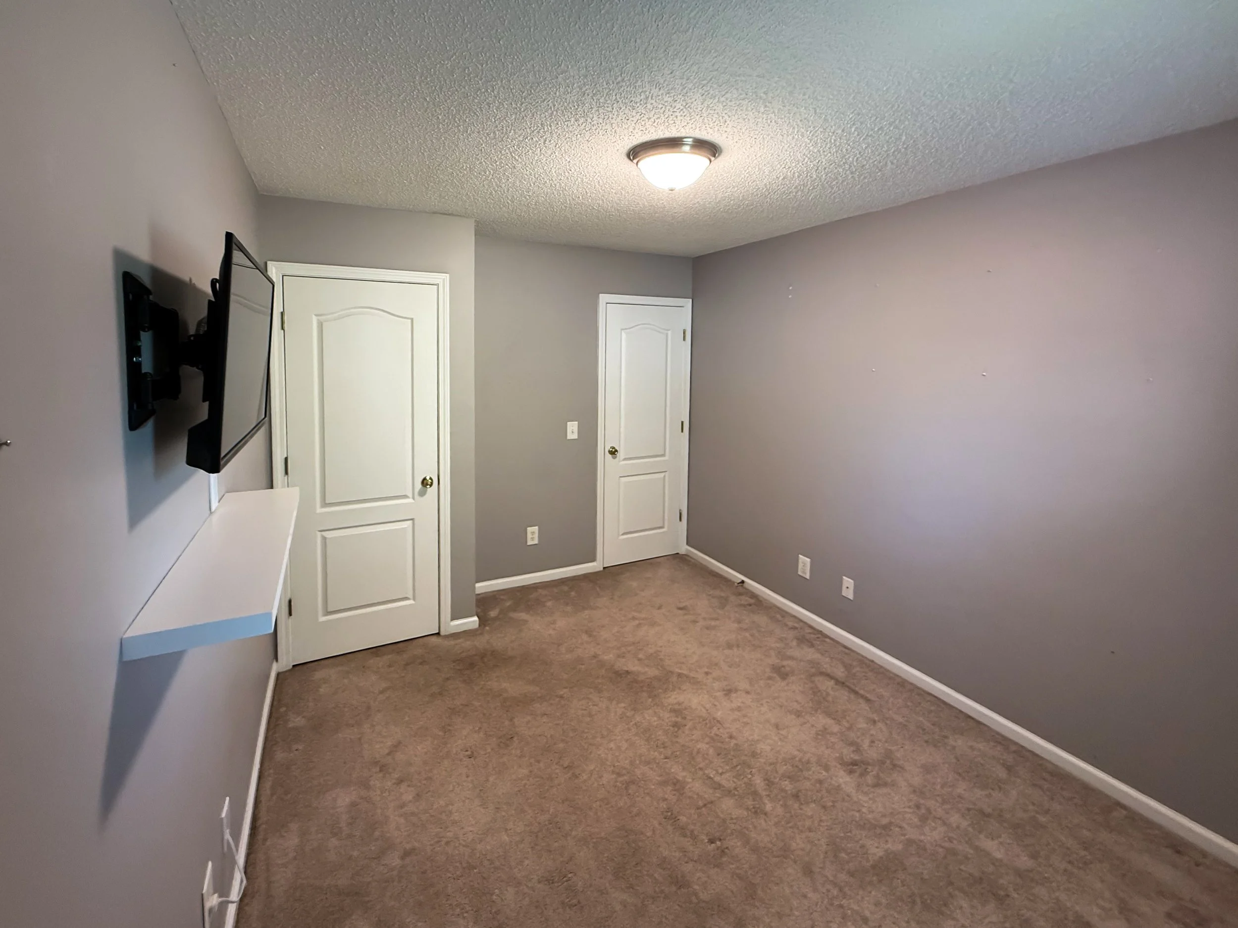 Empty room with gray walls, brown carpeted floor, two closed white doors, wall-mounted TV, and a small white shelf on the wall.