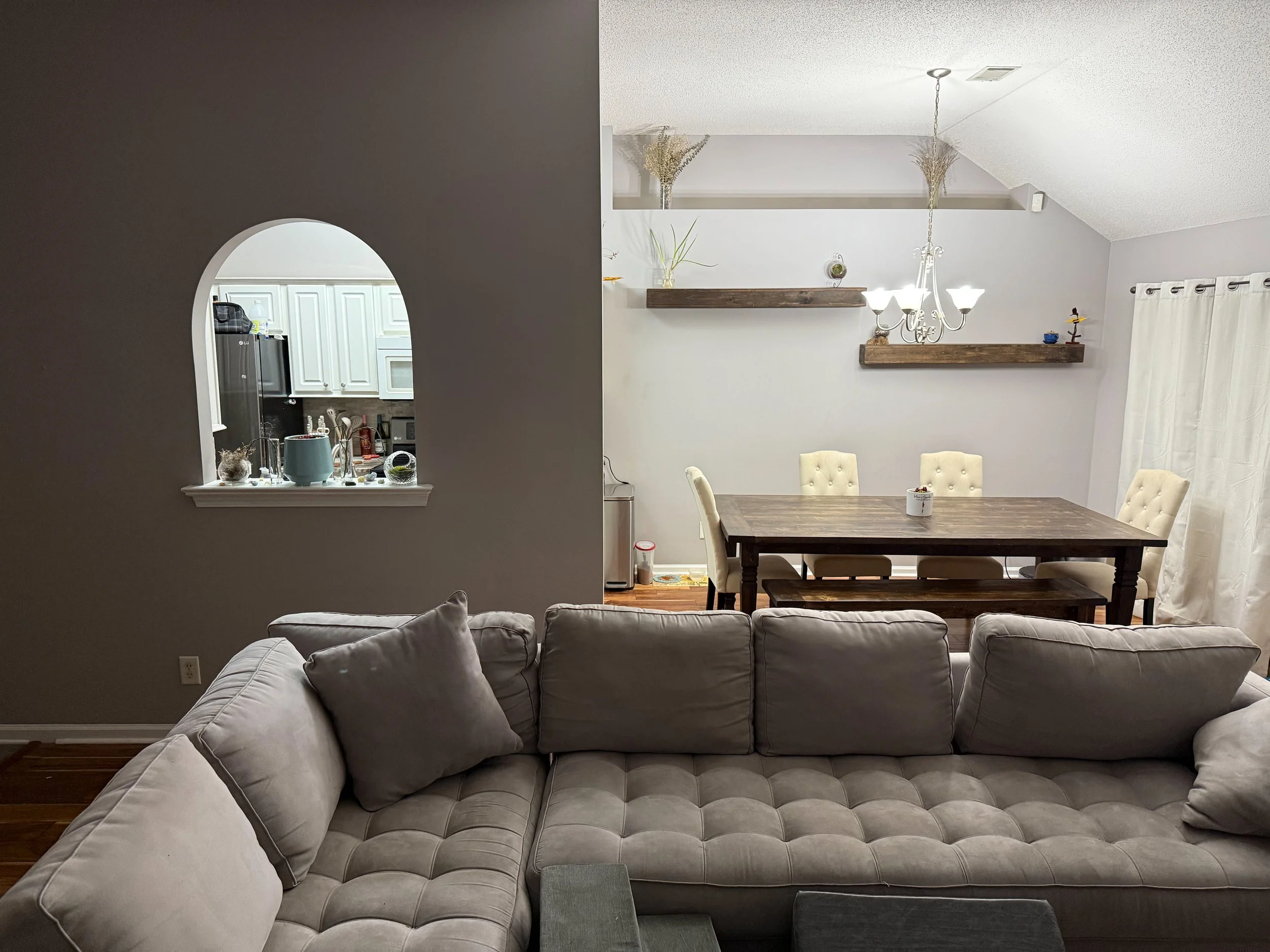 Living room with a beige sofa, a dining table with six white chairs, a chandelier, wall-mounted wooden shelves with decorative items, and a white curtain covering a window. Part of the kitchen with white cabinets and a black refrigerator is visible t