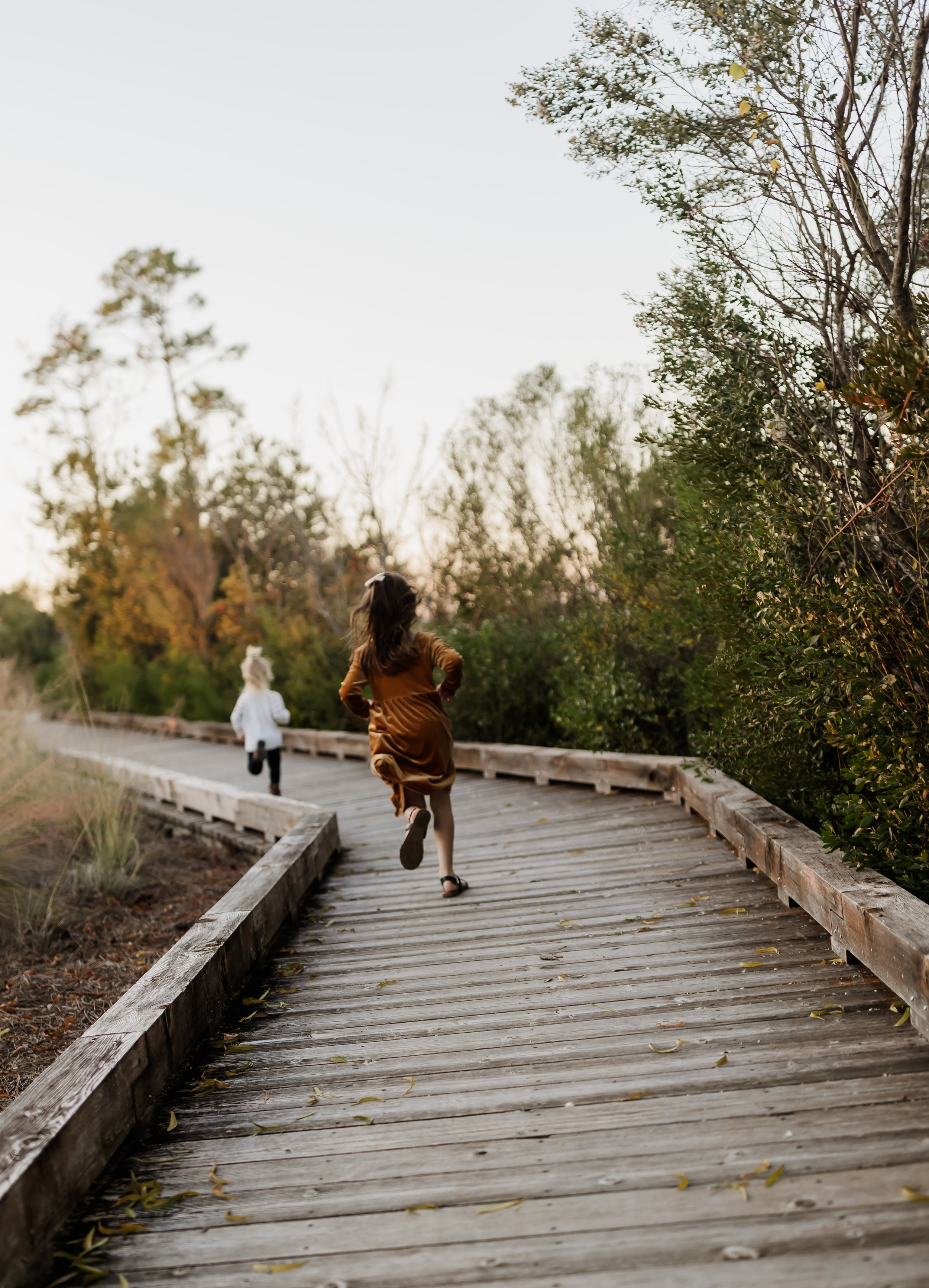 Two children running on a wooden pathway in a natural setting with trees and bushes, during what appears to be late afternoon or early evening.