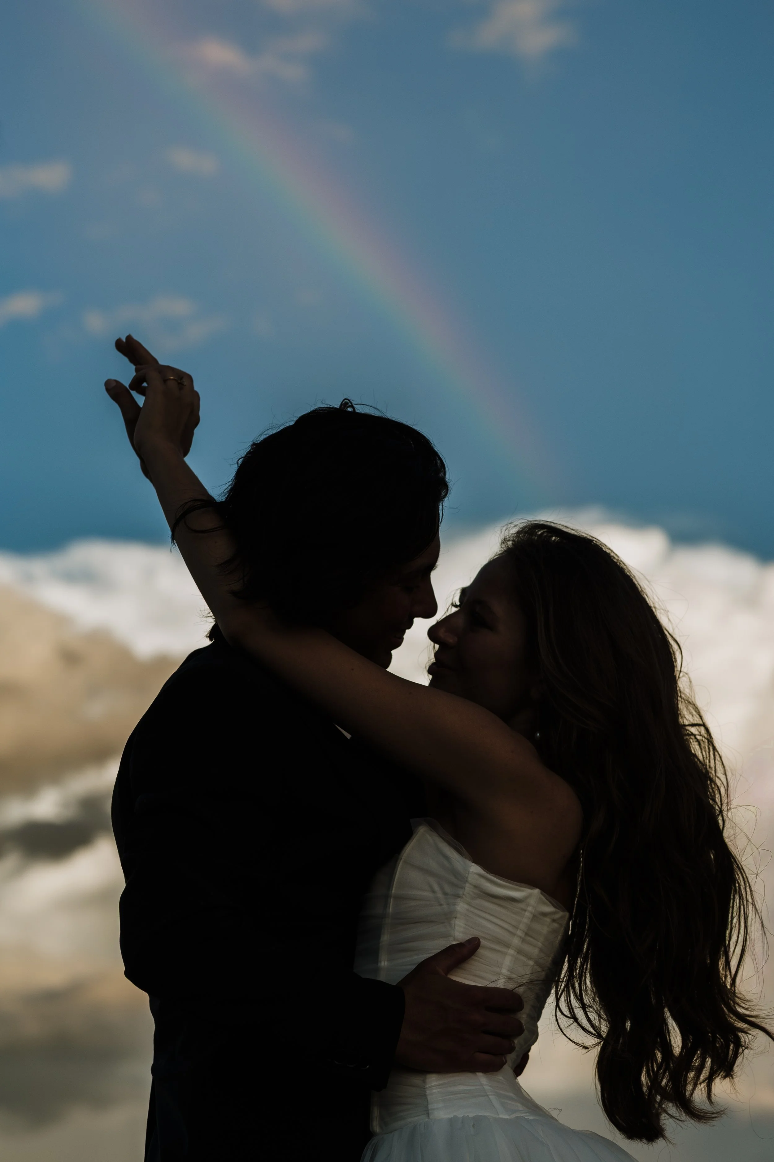 A silhouette of a couple embracing on a beach, with a rainbow in the sky and clouds in the background.