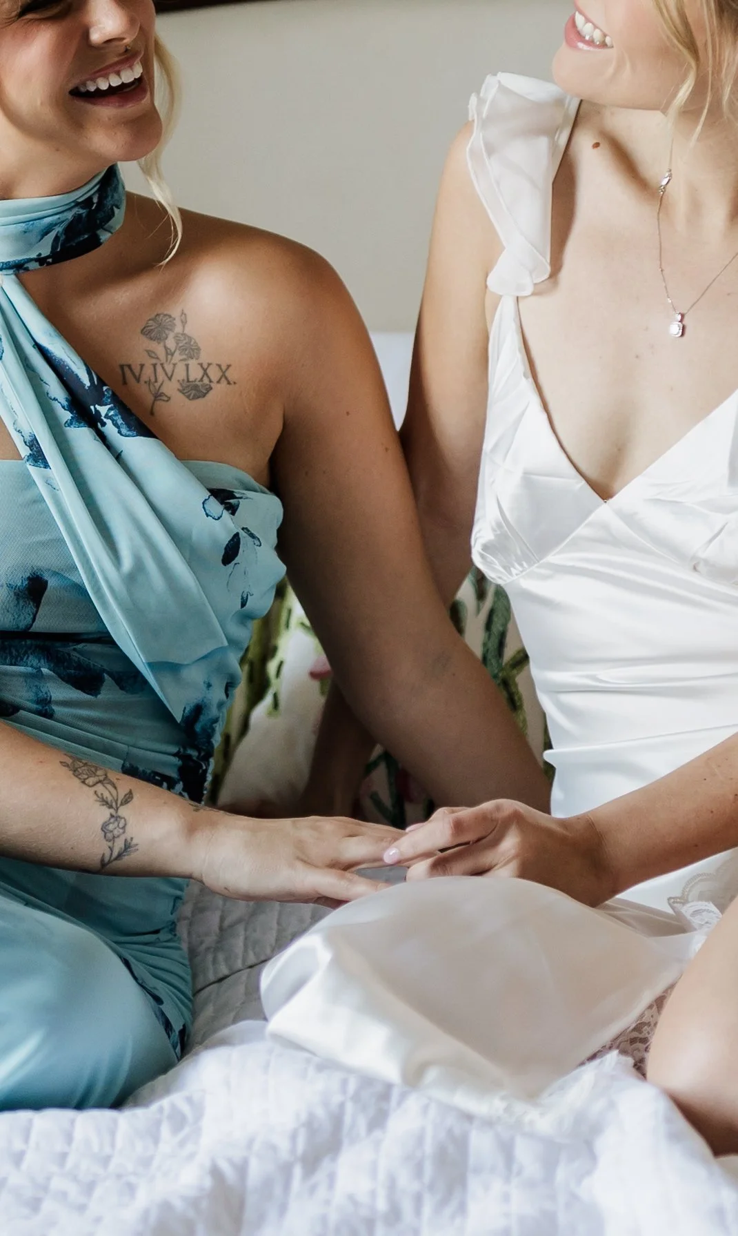 Two women are holding hands and smiling at each other, sitting on a bed. One woman is wearing a blue floral dress and the other is wearing a white dress with ruffled shoulder straps.
