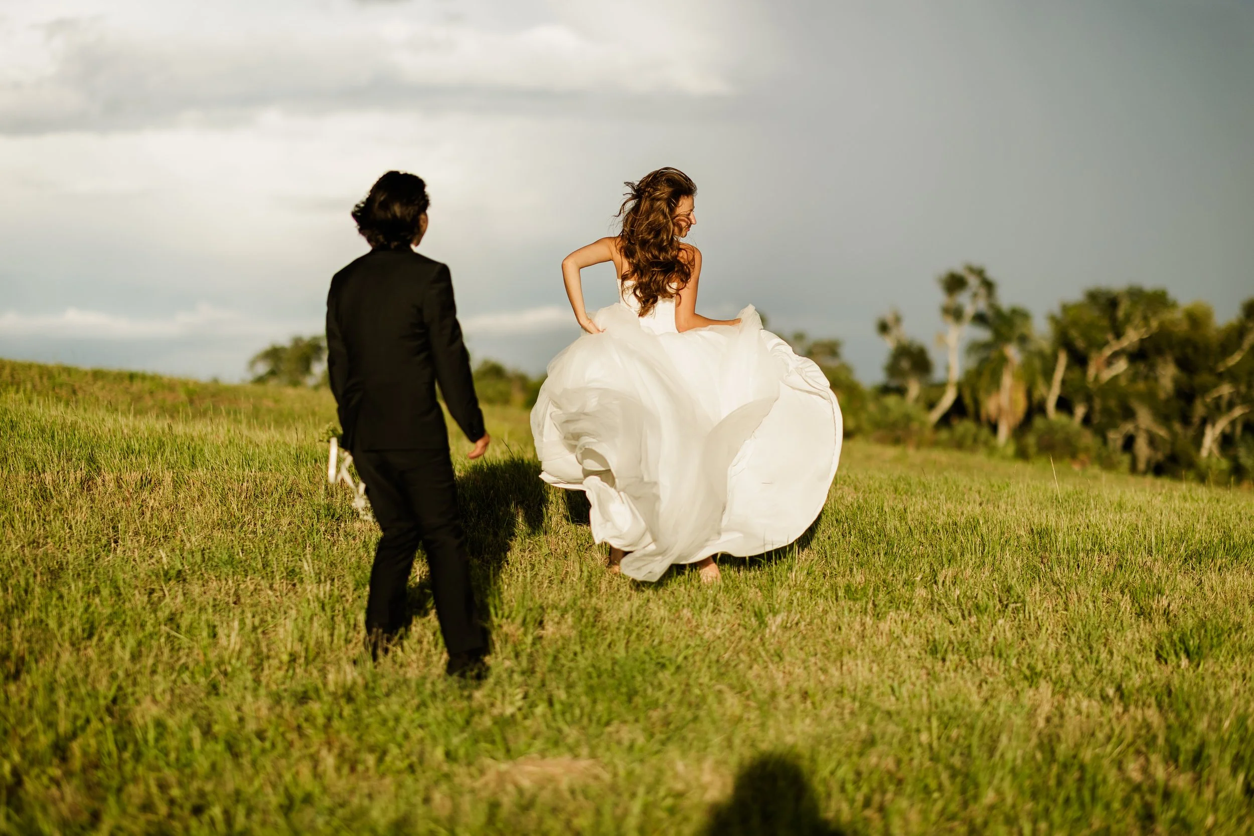 A wedding couple walking in a grassy field, with the bride in a white dress and the groom in a black suit, under a cloudy sky.