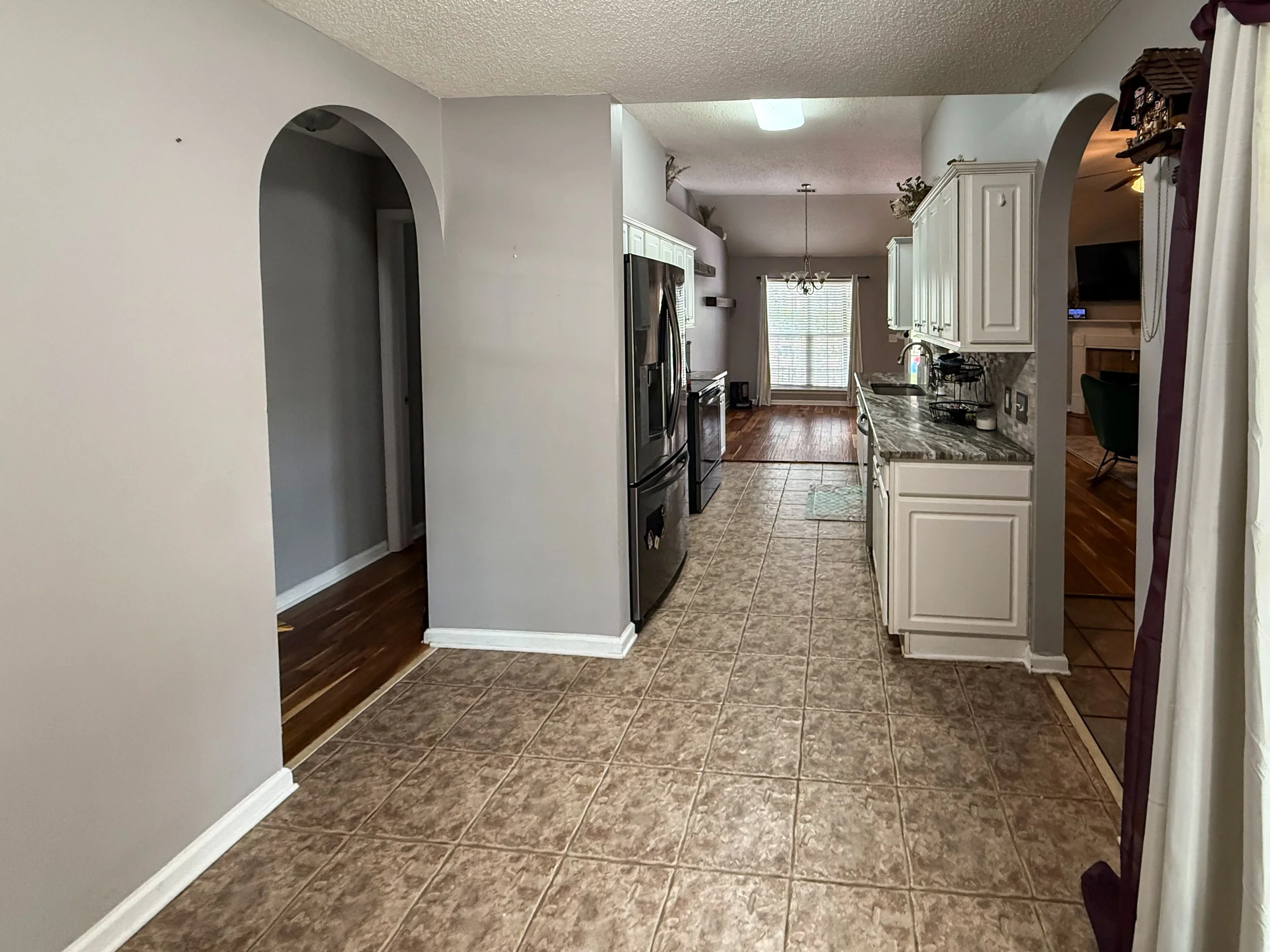 View of a kitchen with white cabinets, stainless steel refrigerator, granite counters, tiled floor, and hardwood flooring in the adjacent room. There is a window with blinds, a chandelier, and a fireplace in the background.