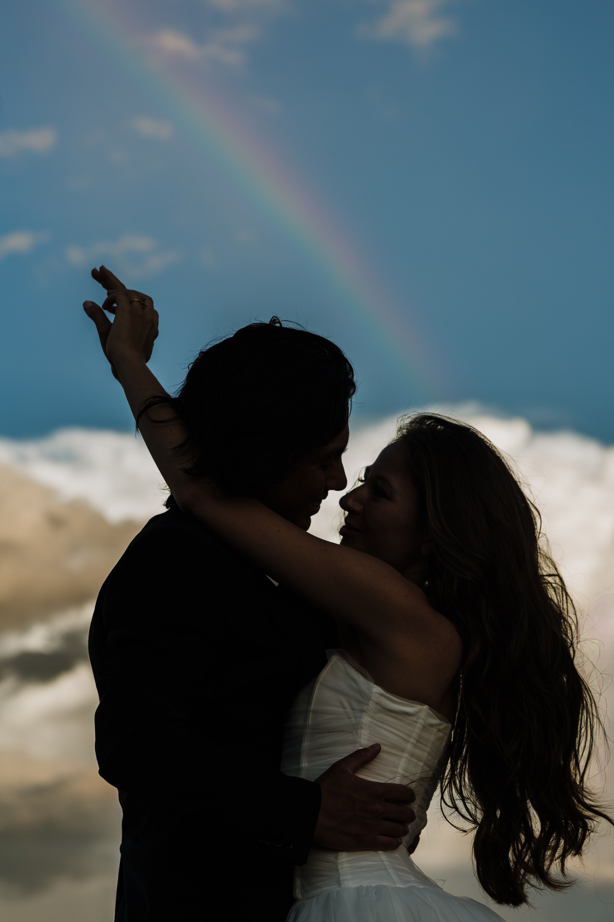 A silhouette of a couple embracing against a background of a partly cloudy sky with a rainbow.