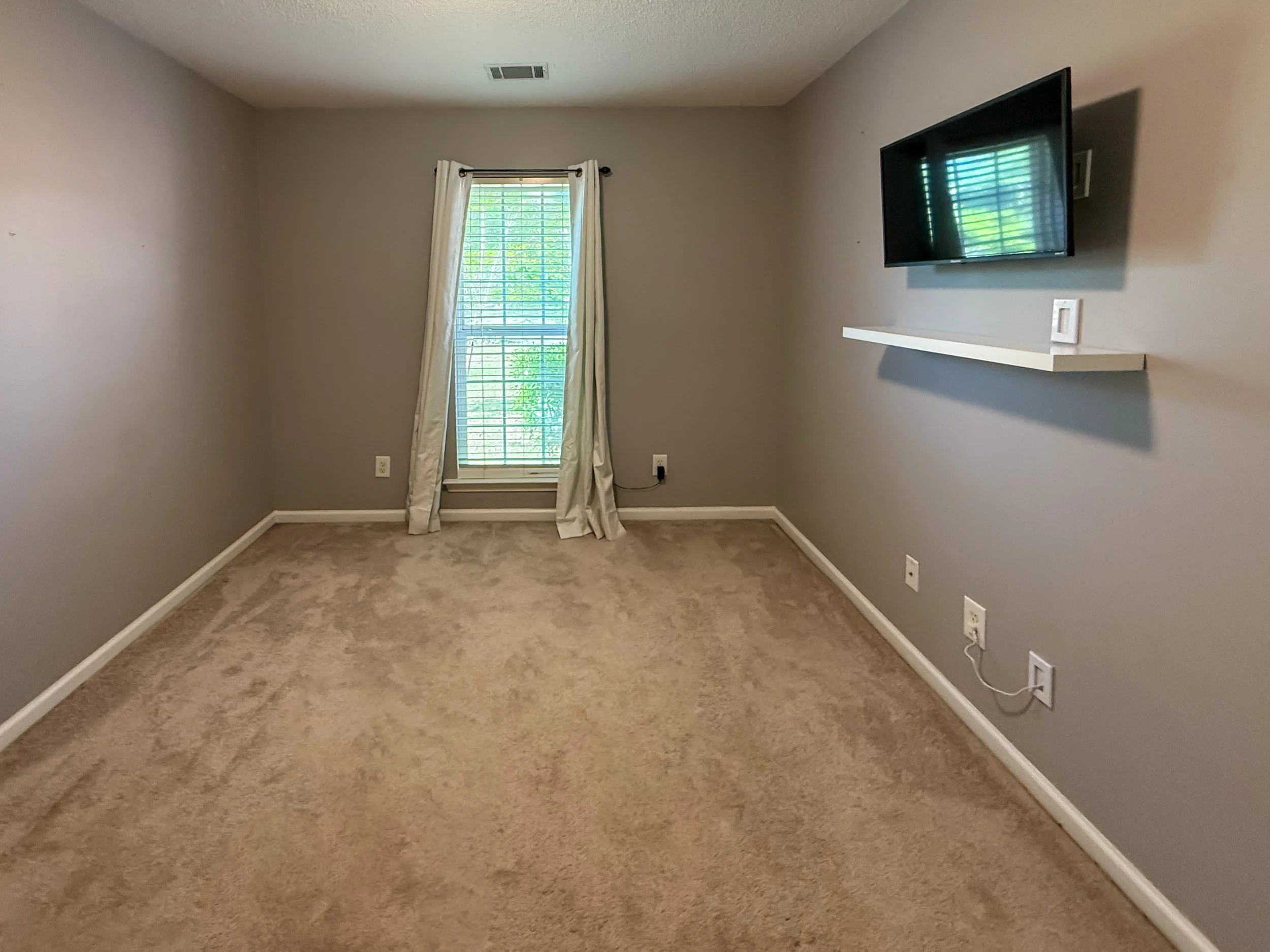 Empty beige-carpeted room with light gray walls, multiple electrical outlets, a window with white curtains, and a wall-mounted television with a small floating shelf underneath.