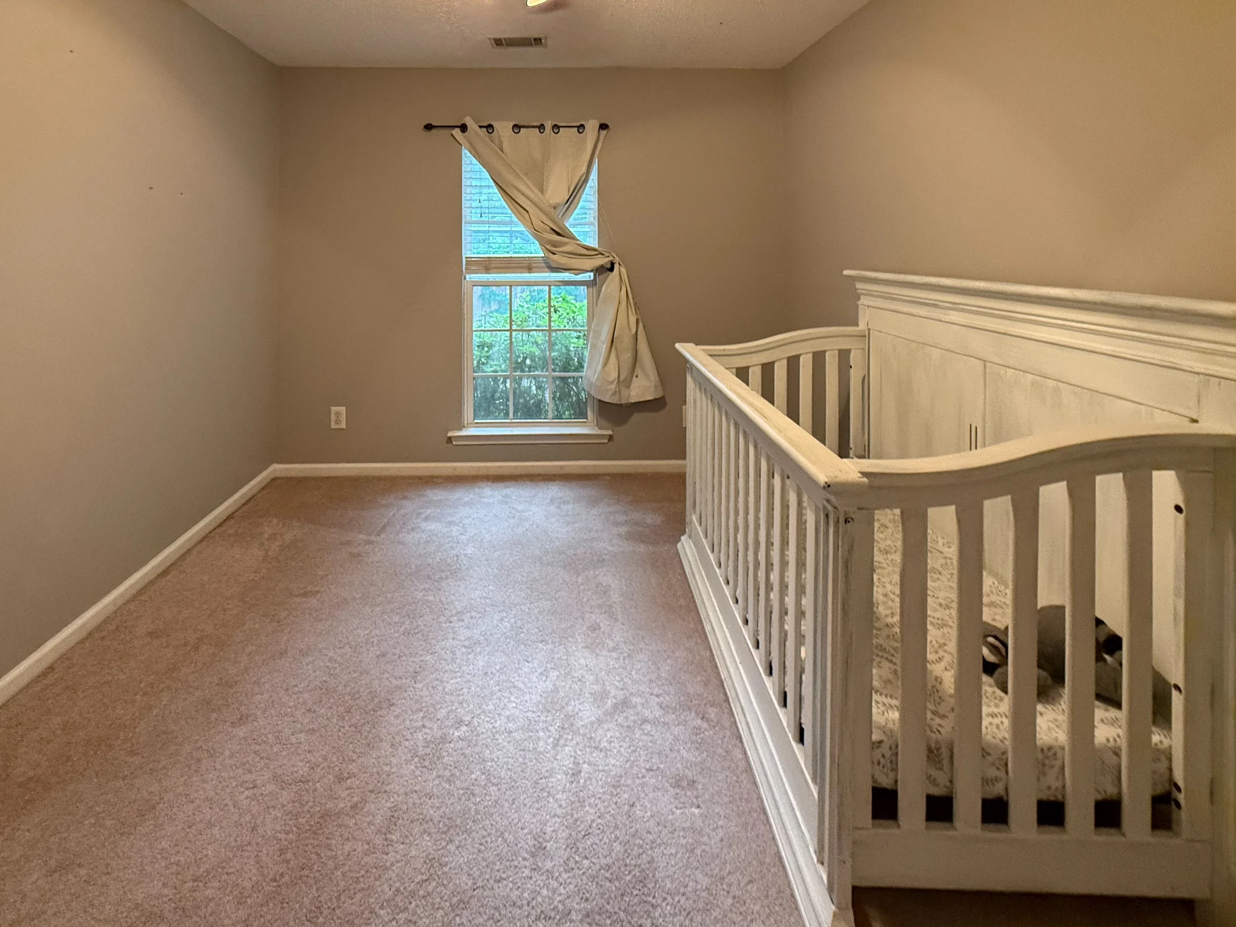 Empty room with beige walls, a window with partially open white curtains, and a white wooden railing/partition.