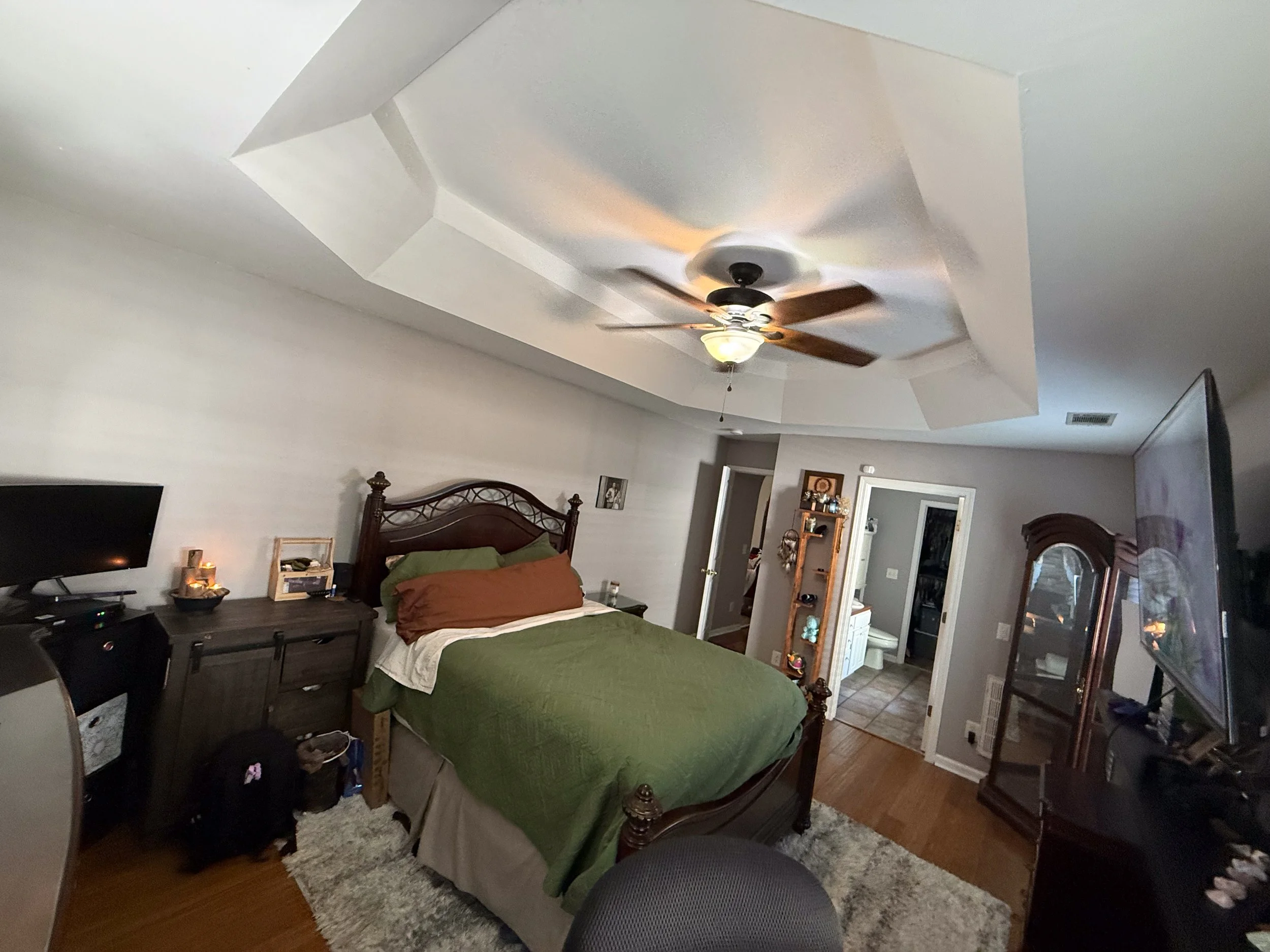 A bedroom with a wooden bed frame and green bedding, a nightstand with candles, a dresser, a TV, and a ceiling fan with wooden blades.
