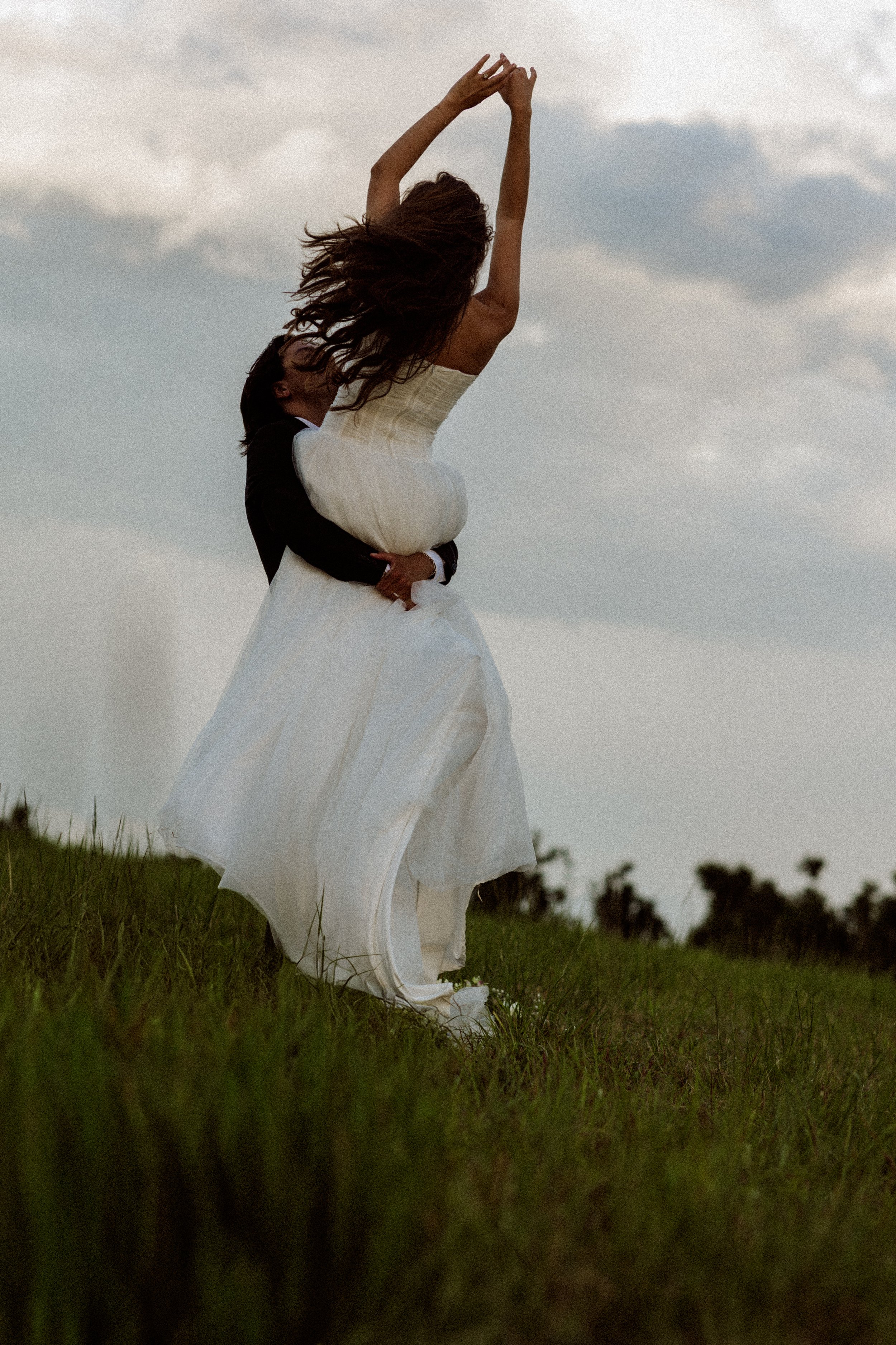A couple dancing outdoors on a grassy field under a cloudy sky, with the woman wearing a white dress and the man in a suit.