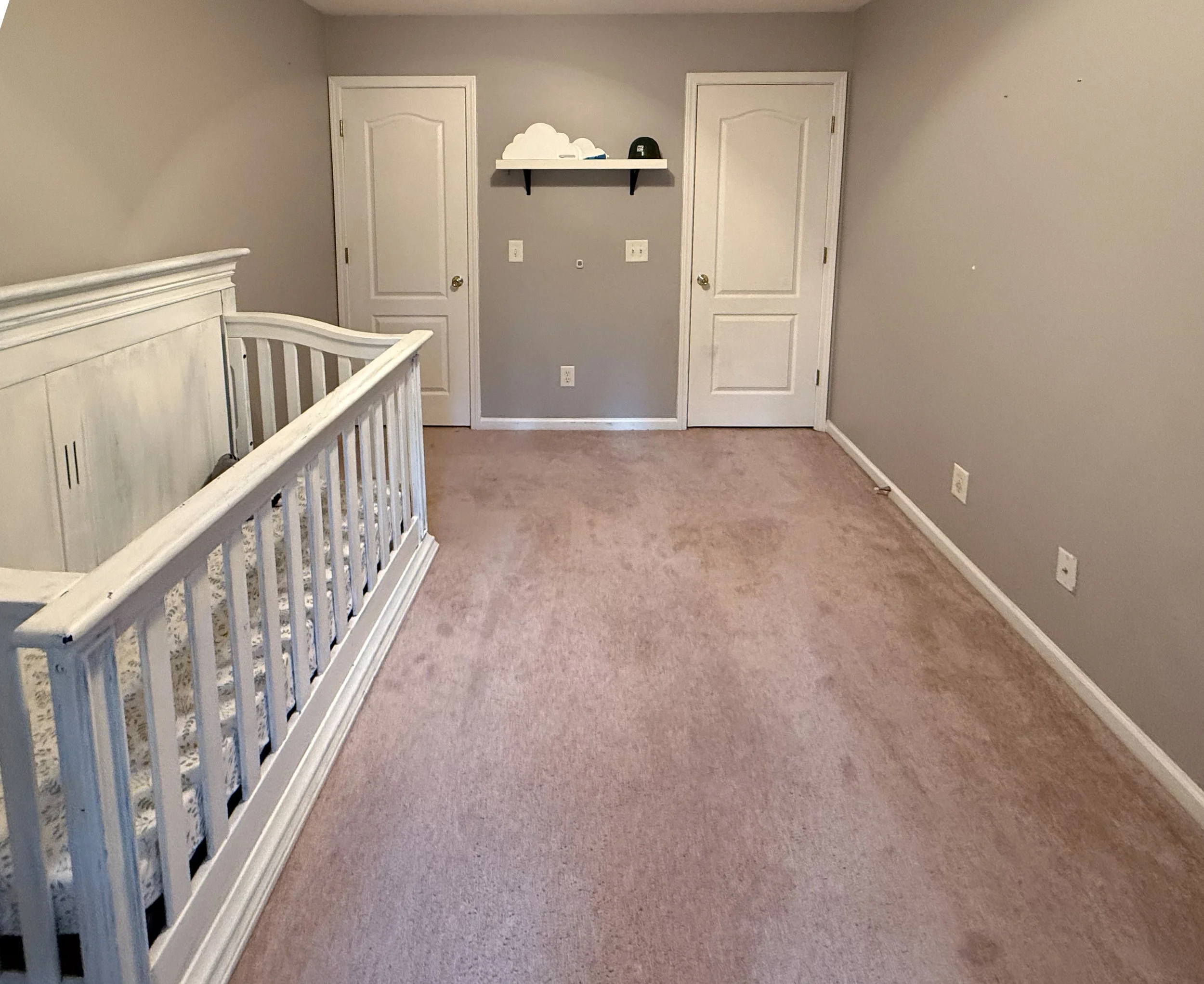 Empty nursery with a white crib, gray walls, a small floating shelf with a cloud decoration, a black cap, and two doors.
