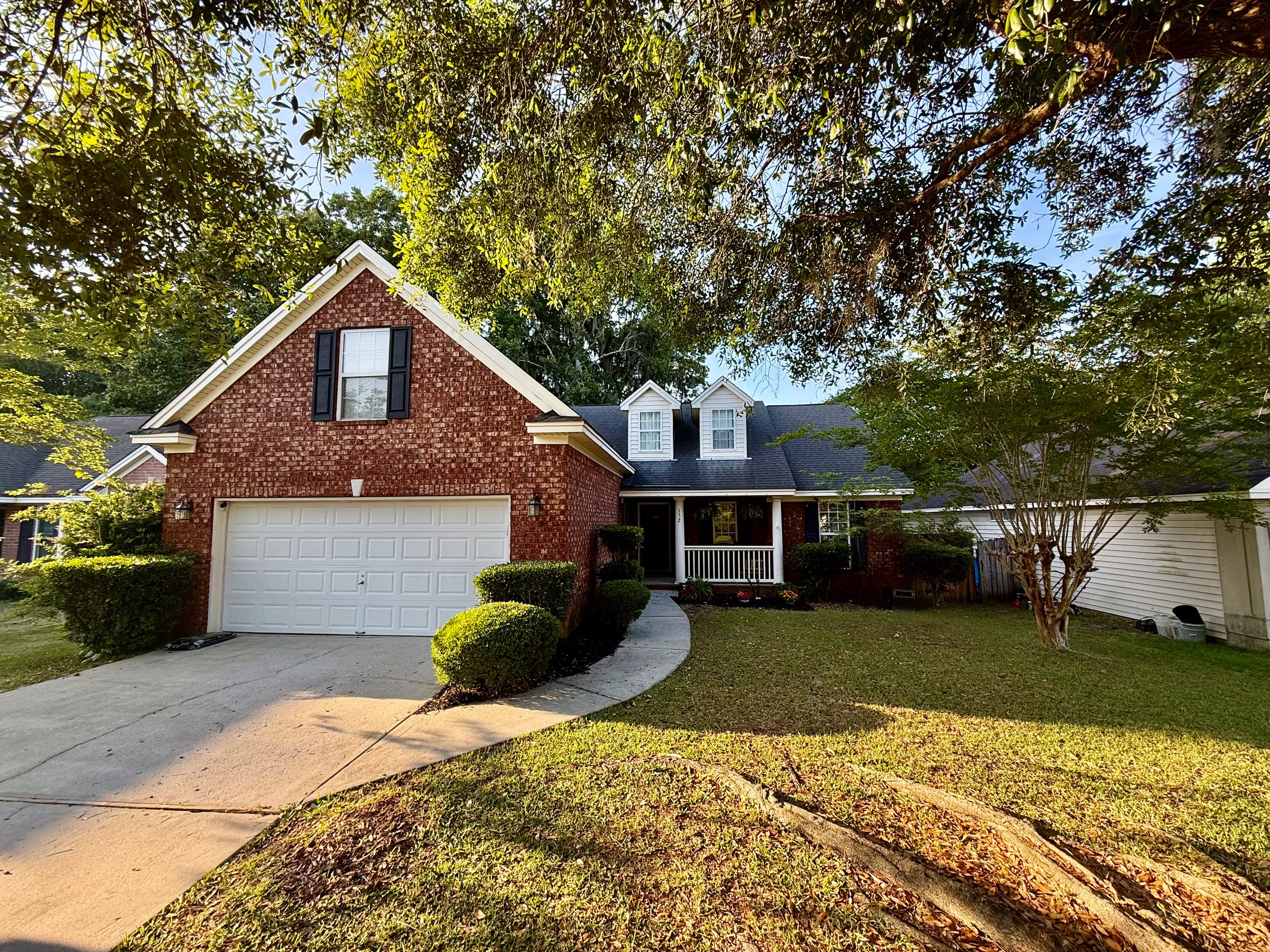 A red brick house with a white garage door, dark shutters, and a front porch, surrounded by trees and bushes on a sunny day.