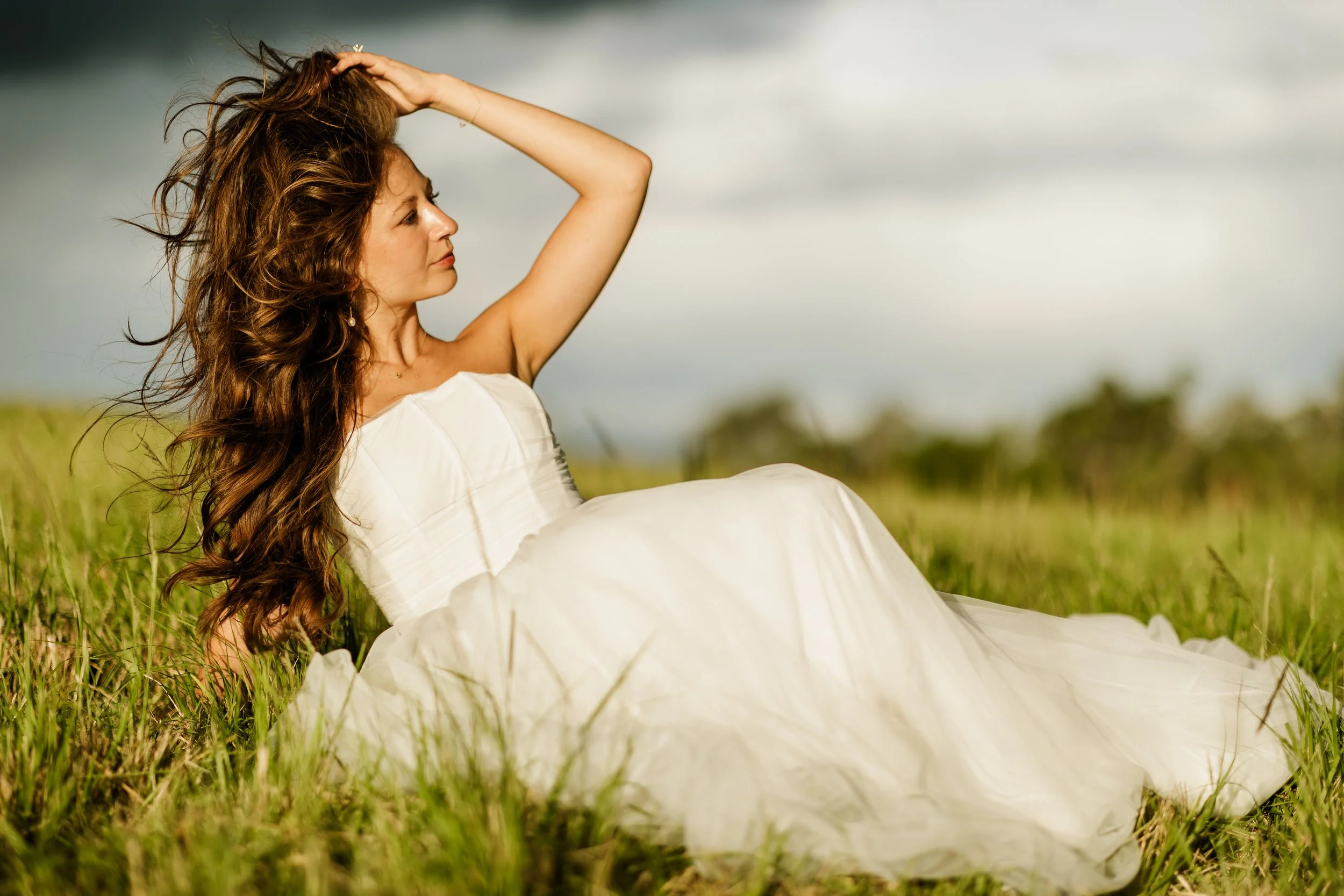 A woman with long, wavy brown hair sitting in a grassy field on a cloudy day, wearing a white dress, looking to the side with her hand on her head.