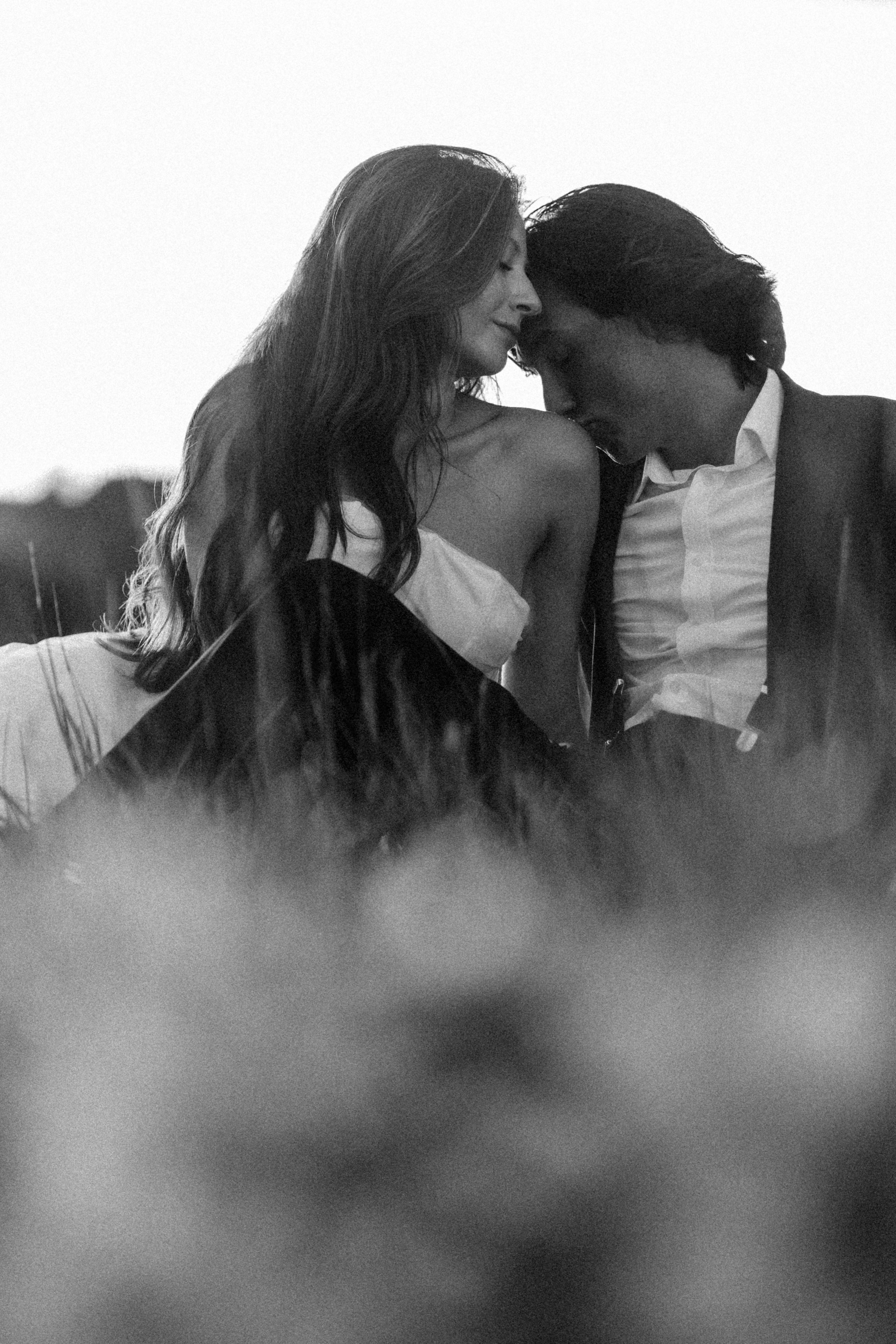 Black and white photo of a man and woman with their foreheads touching and eyes closed, sharing an intimate moment outdoors.