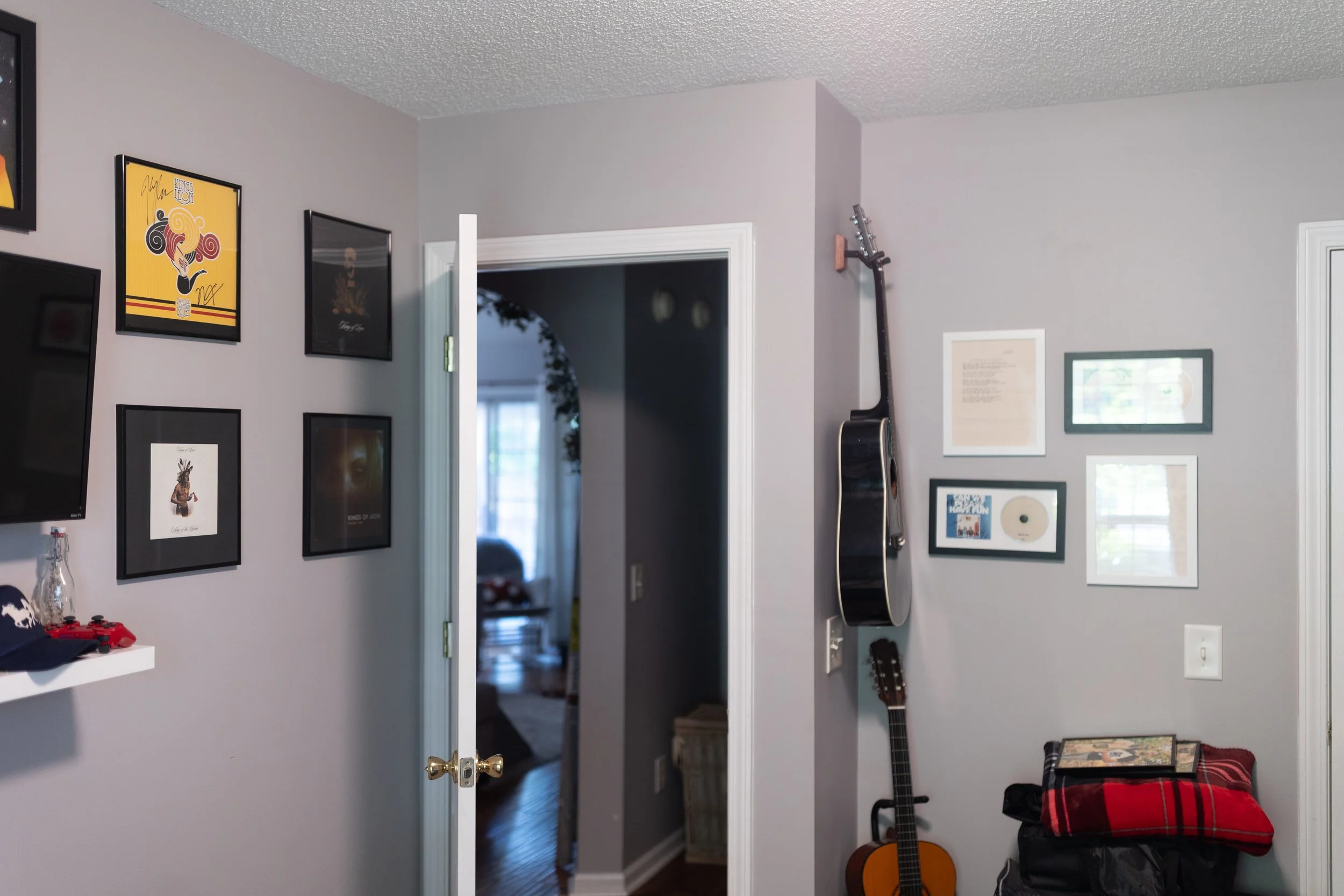 A view of a room with light gray walls decorated with framed artwork, a guitar leaning against the wall, and a doorway leading to another room. There is a guitar on a stand, a stereo, and a red plaid blanket on a chair.