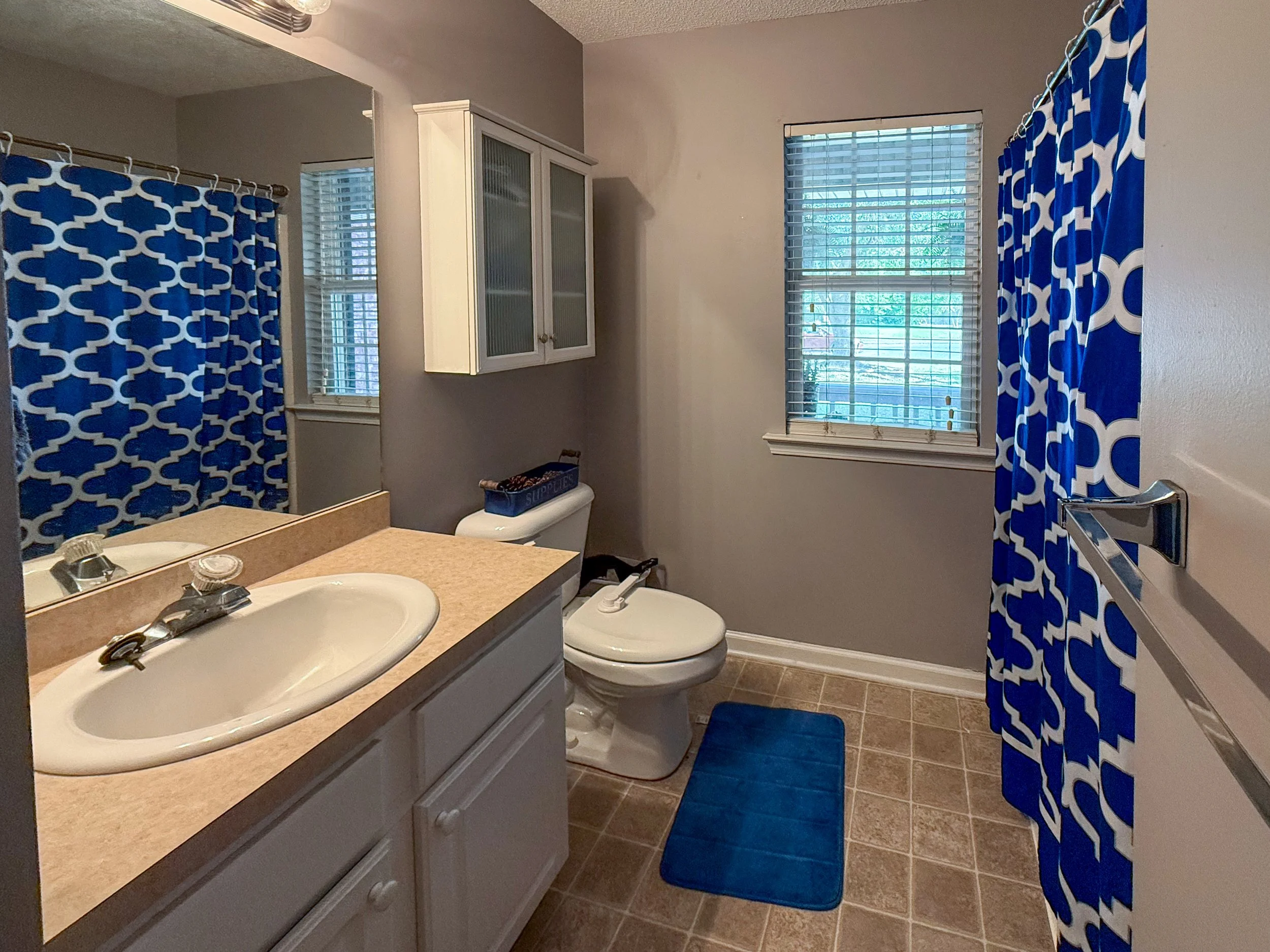 Bathroom with two sinks, a toilet, a window with blinds, blue patterned shower curtain, and a white wall cabinet.