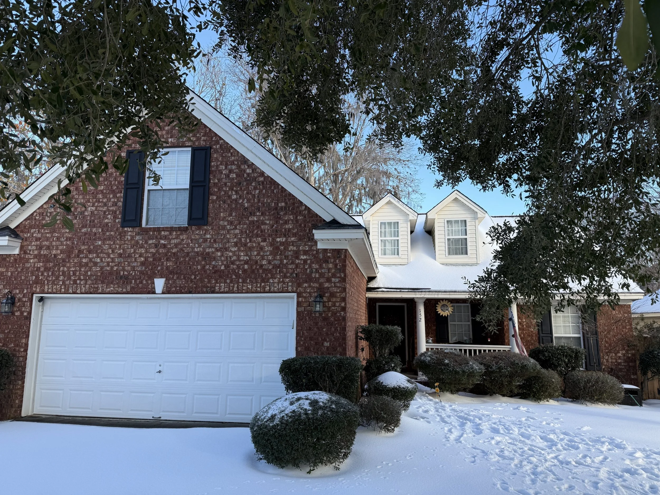 A suburban house with a brick facade, white garage door, snow-covered front yard, and trees with snow on their branches during winter.
