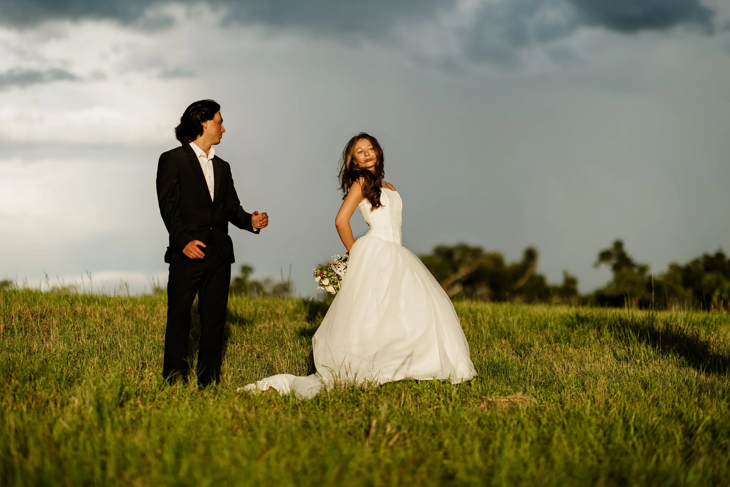 A man in a black suit and a woman in a white wedding dress standing outdoors on a grassy field under a cloudy sky.