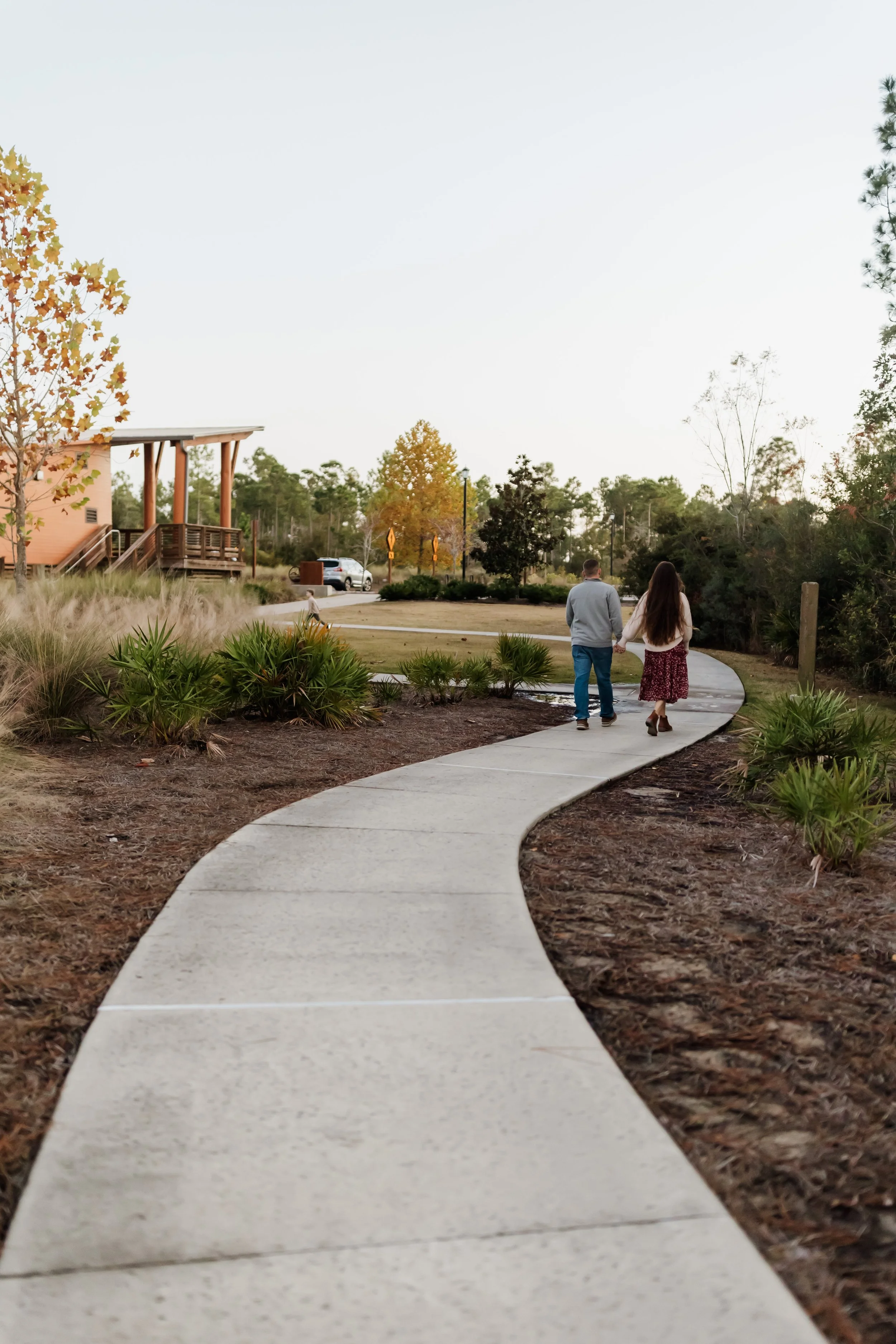 A curved concrete sidewalk in a park with trees and shrubs, a couple walking hand in hand, and a building with stairs in the background during late afternoon or early evening.