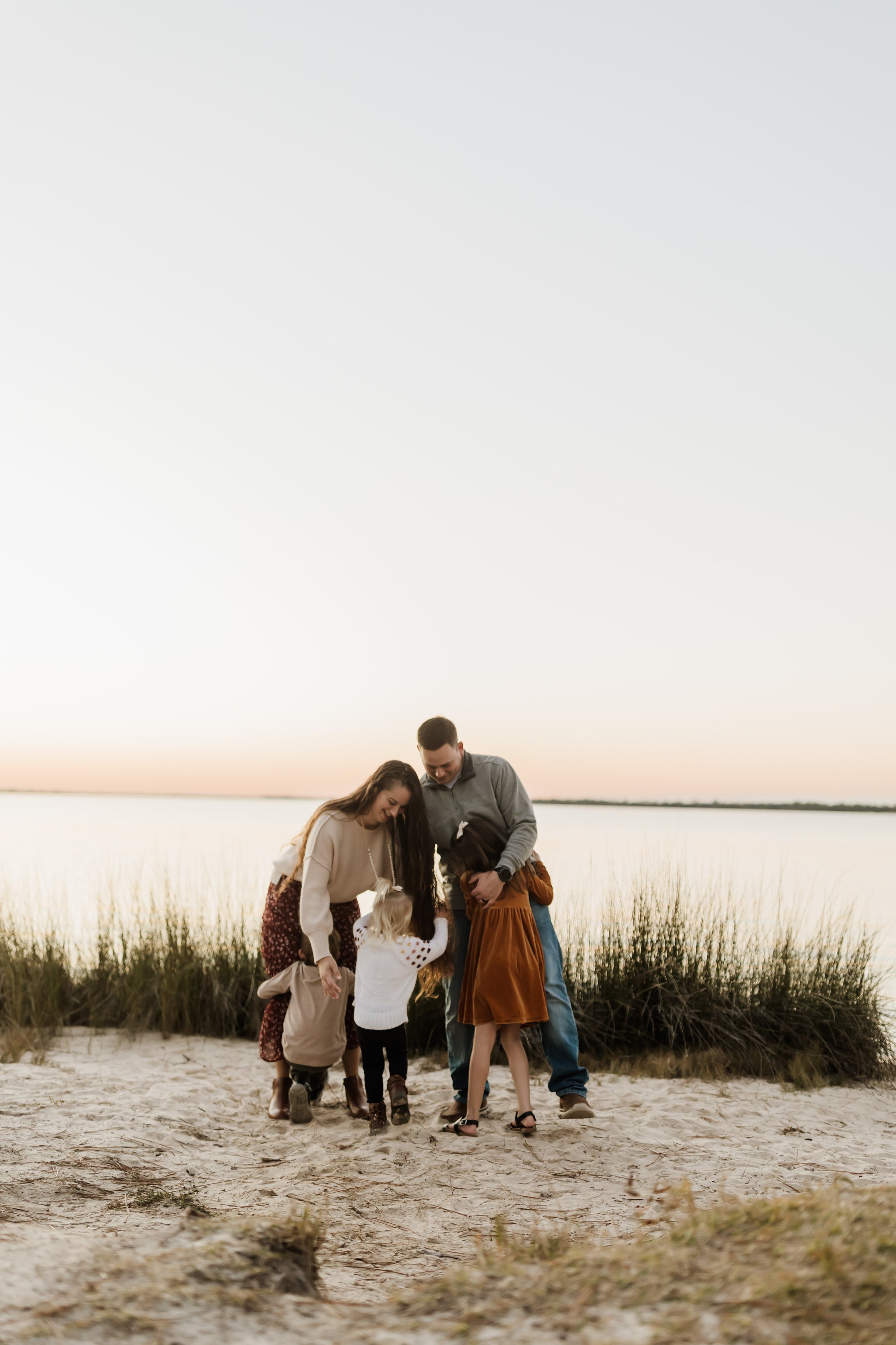 A family of five, including three children, standing on a sandy beach at sunset, examining something on the ground.