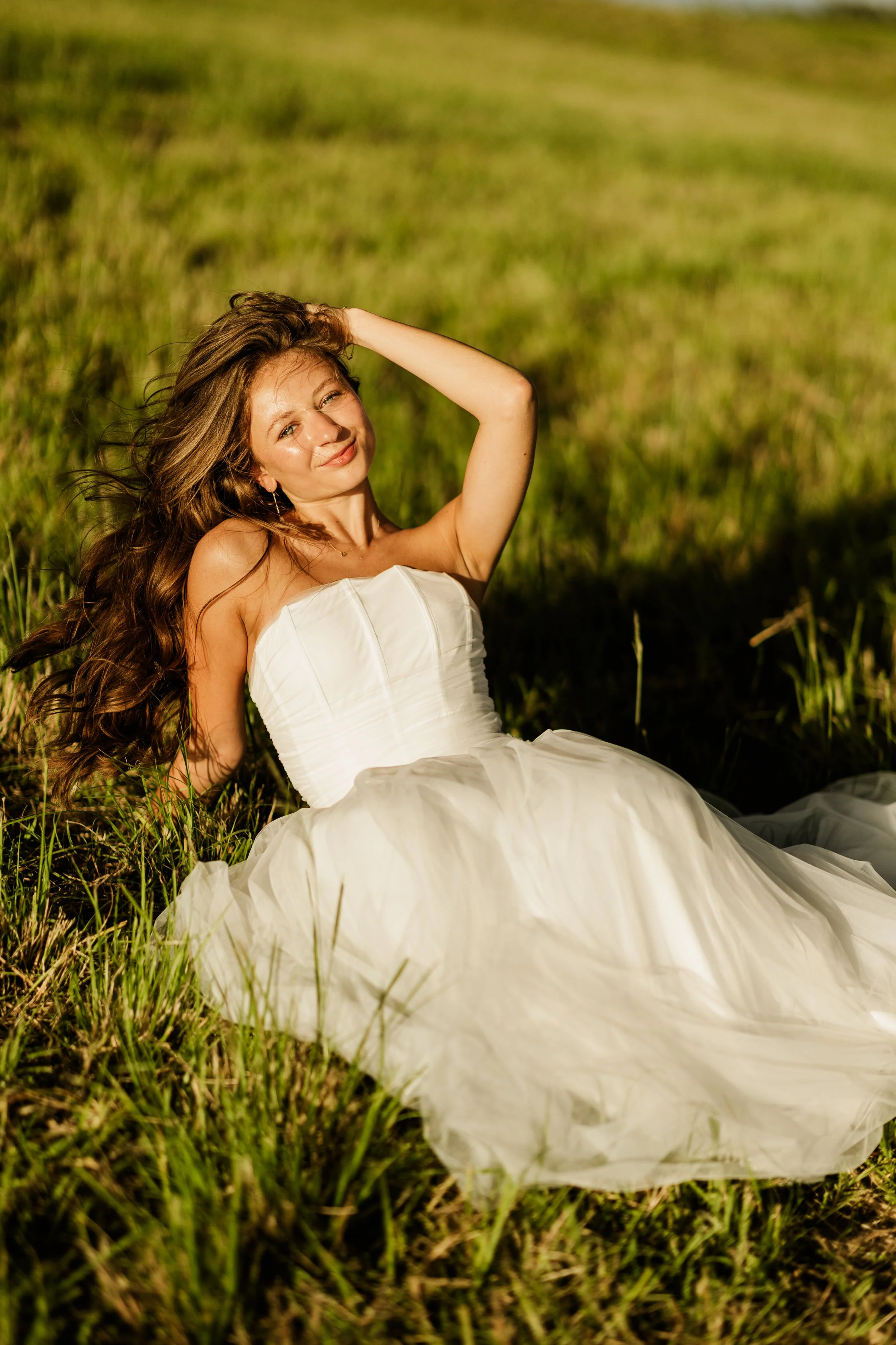 A woman in a white dress sitting in a grassy field on a sunny day, smiling and looking into the camera.