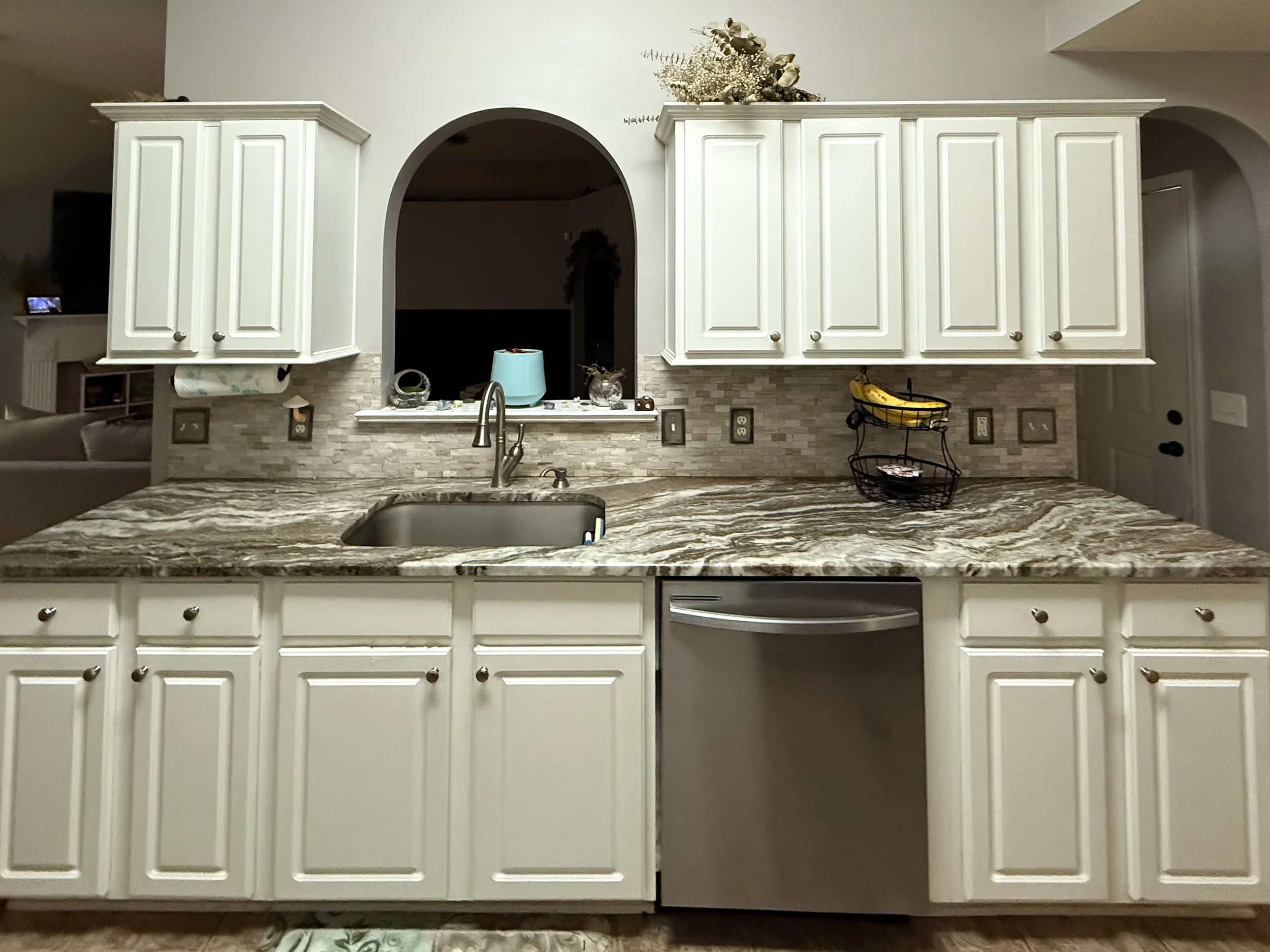 Home kitchen with white cabinets, granite countertop, and a tiled backsplash. Items on the counter include a blue container, a basket of bananas, and decorative items. An arched opening leads to another room.