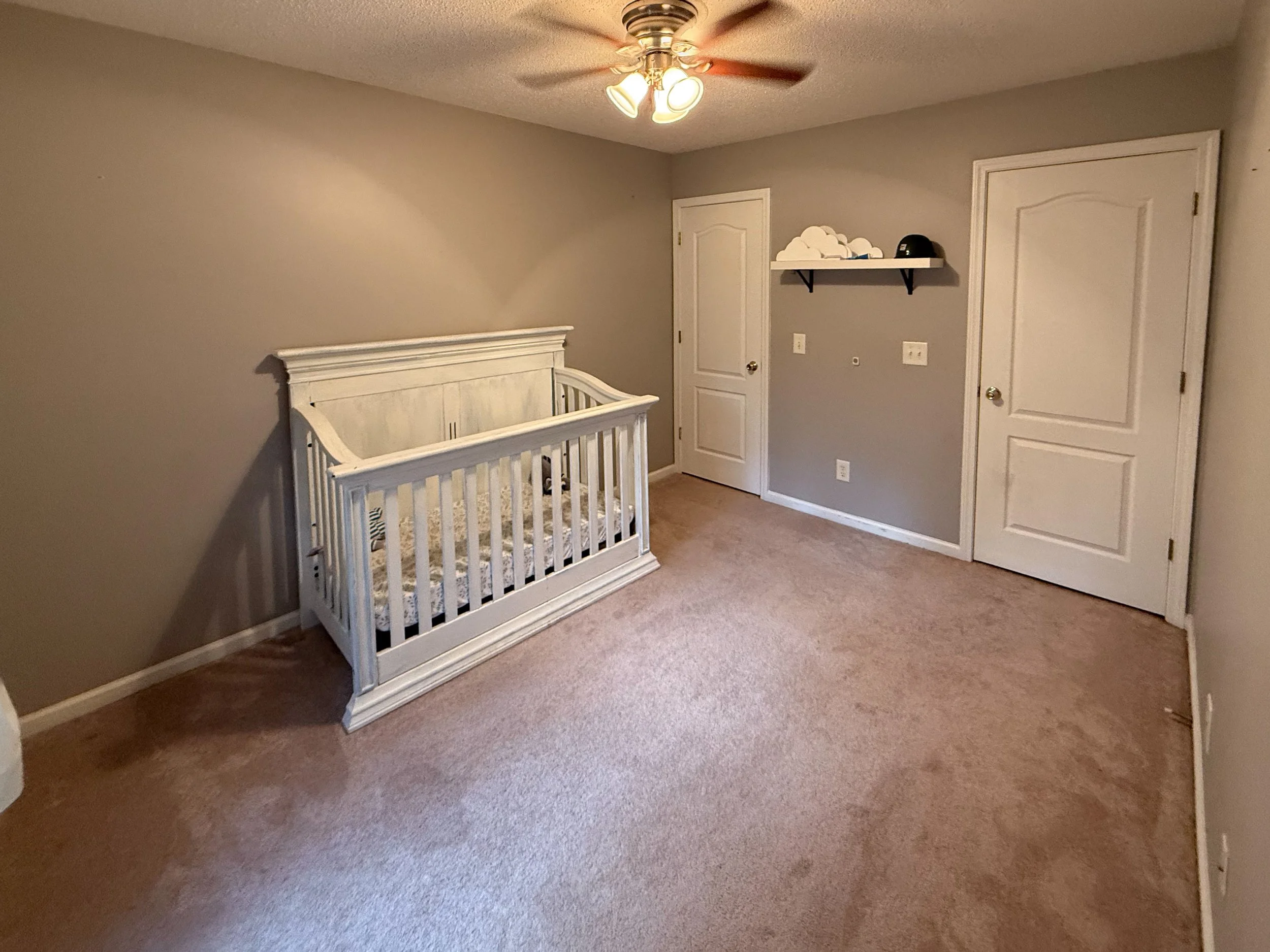 A bedroom with beige carpet, gray walls, a white wooden crib, a white shelf with cloud-shaped decor, a black helmet, and a ceiling fan with lights turned on.