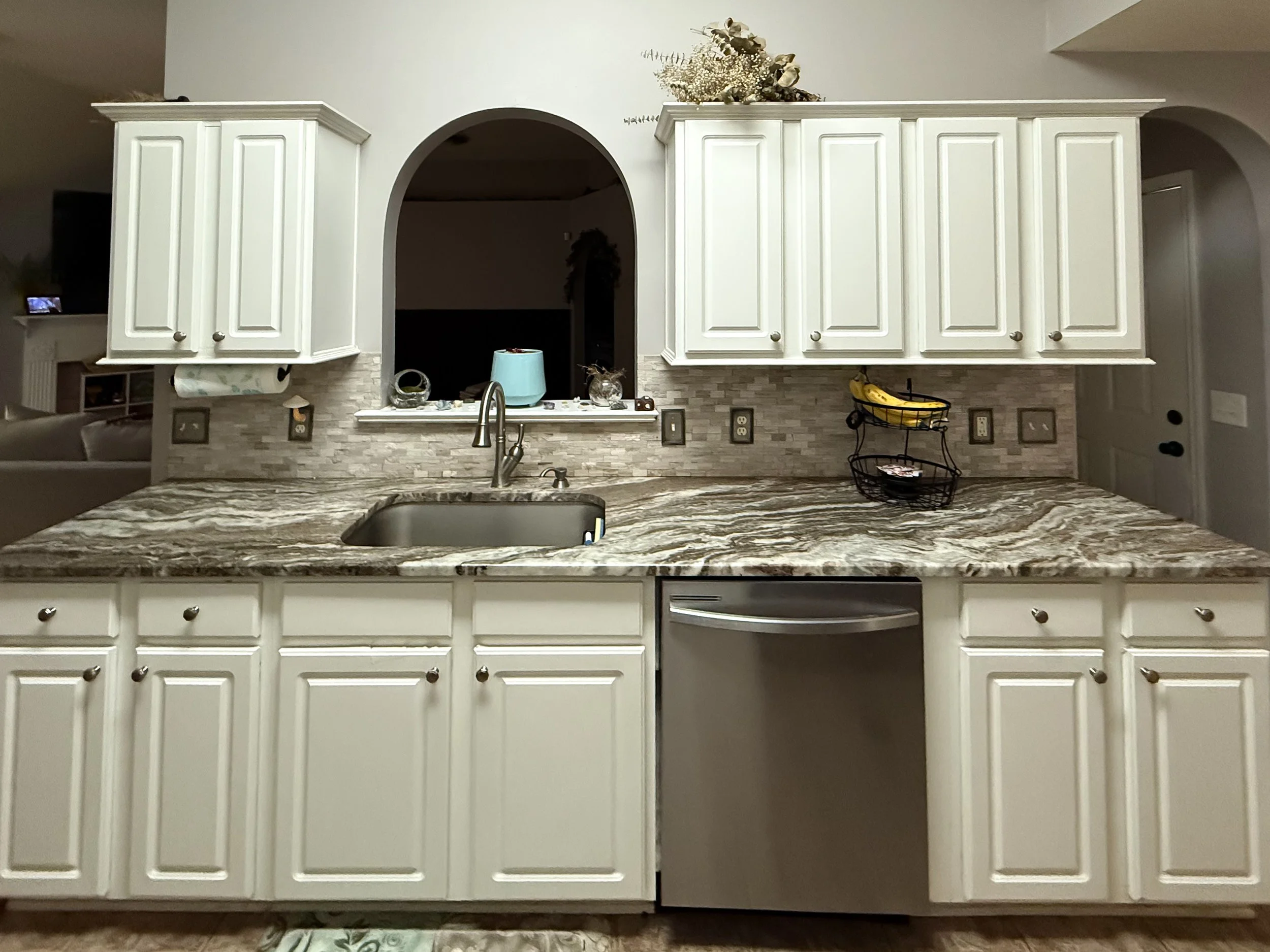 Panoramic view of a kitchen island with a granite countertop, white cabinets, and a stainless steel sink, with a backsplash of beige textured tiles. There are various items on the countertop, including a wire fruit basket with bananas, and some decor