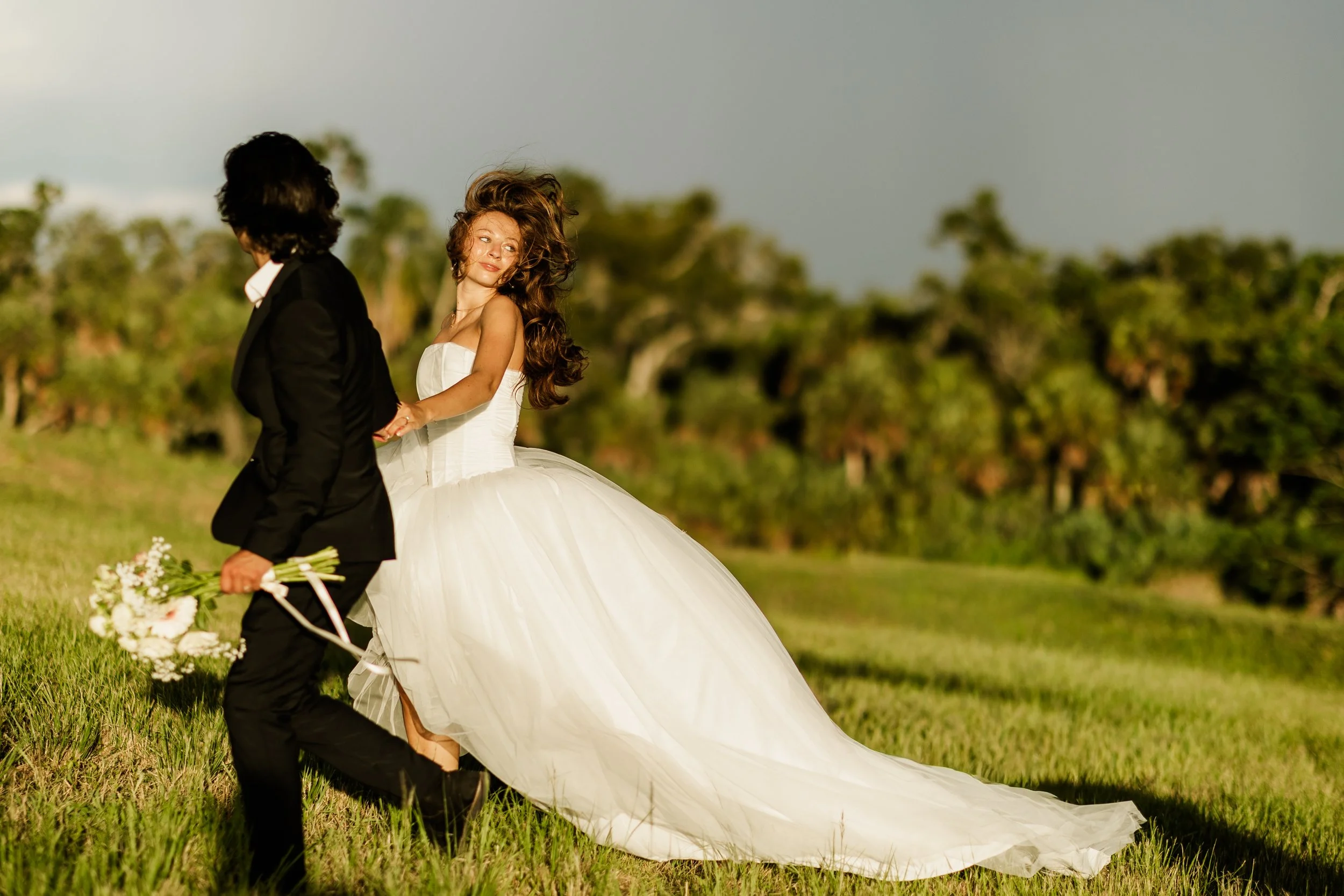 A bride in a white wedding dress and a groom in a black suit holding hands outdoors on a grassy field, with trees in the background.