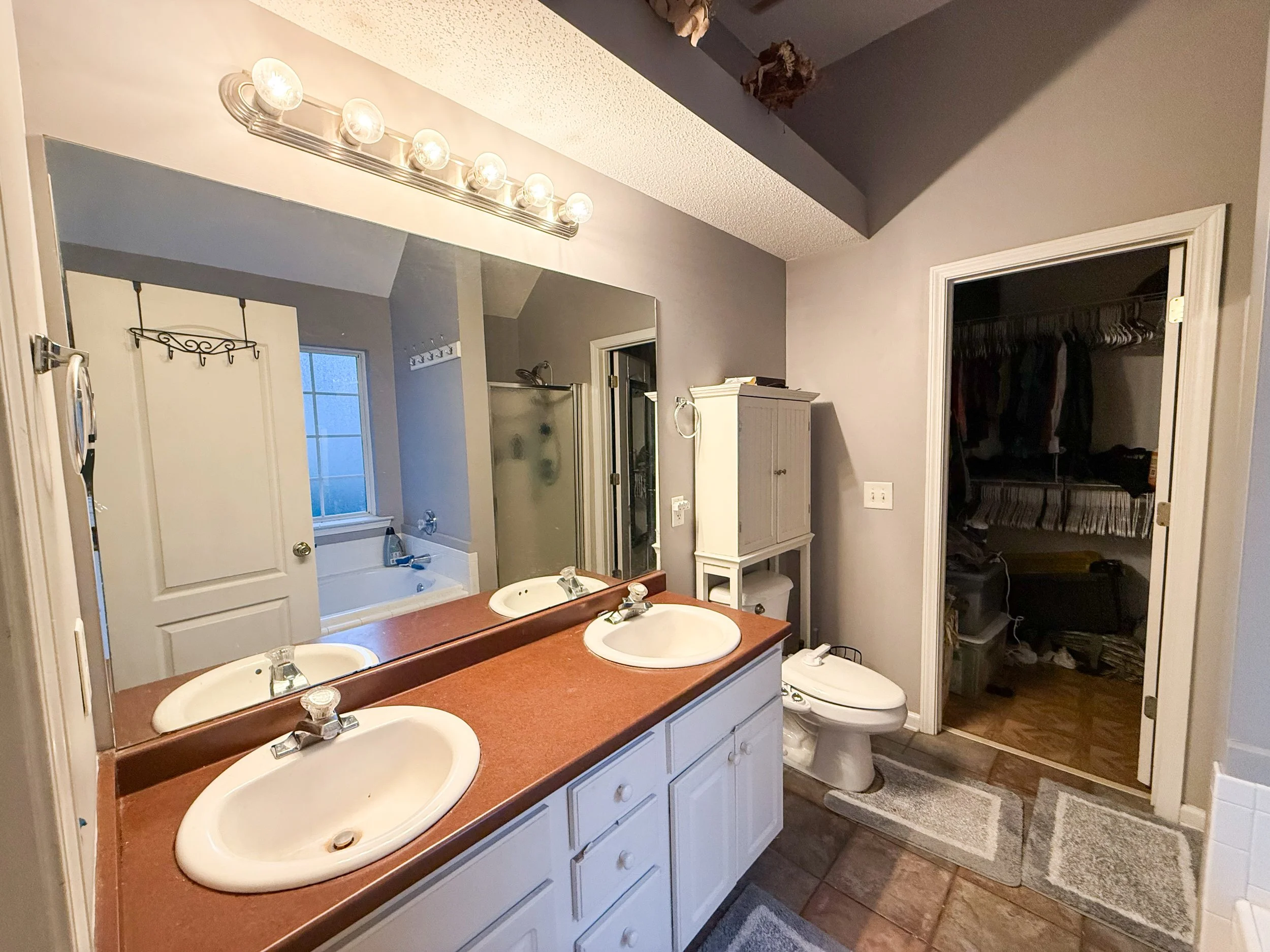 Bathroom with a double sink vanity, large mirror, and a light fixture with five bulbs. There's a window and a bathtub in the background, and an open closet with clothes on hangers. The bathroom has gray walls and brown flooring.