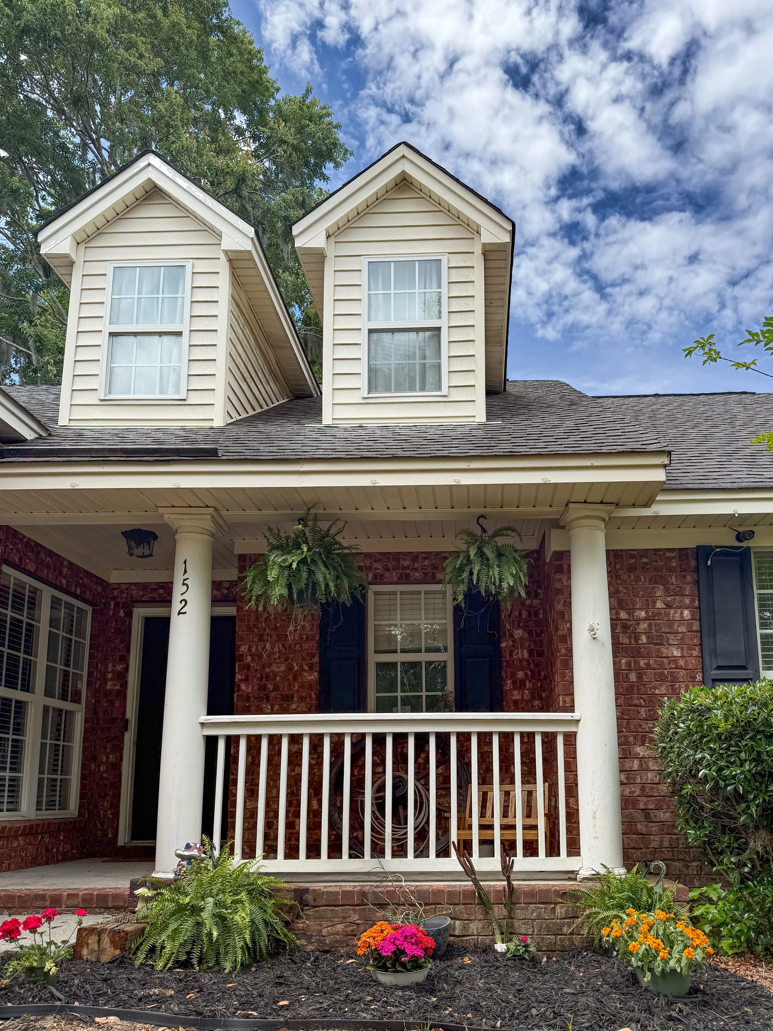 Front view of a house with red brick exterior, white columns, and shutters. Two small dormer windows with white frames and beige siding. The porch has a white railing and hanging plants. Flower beds with pink, orange, and purple flowers, and various 