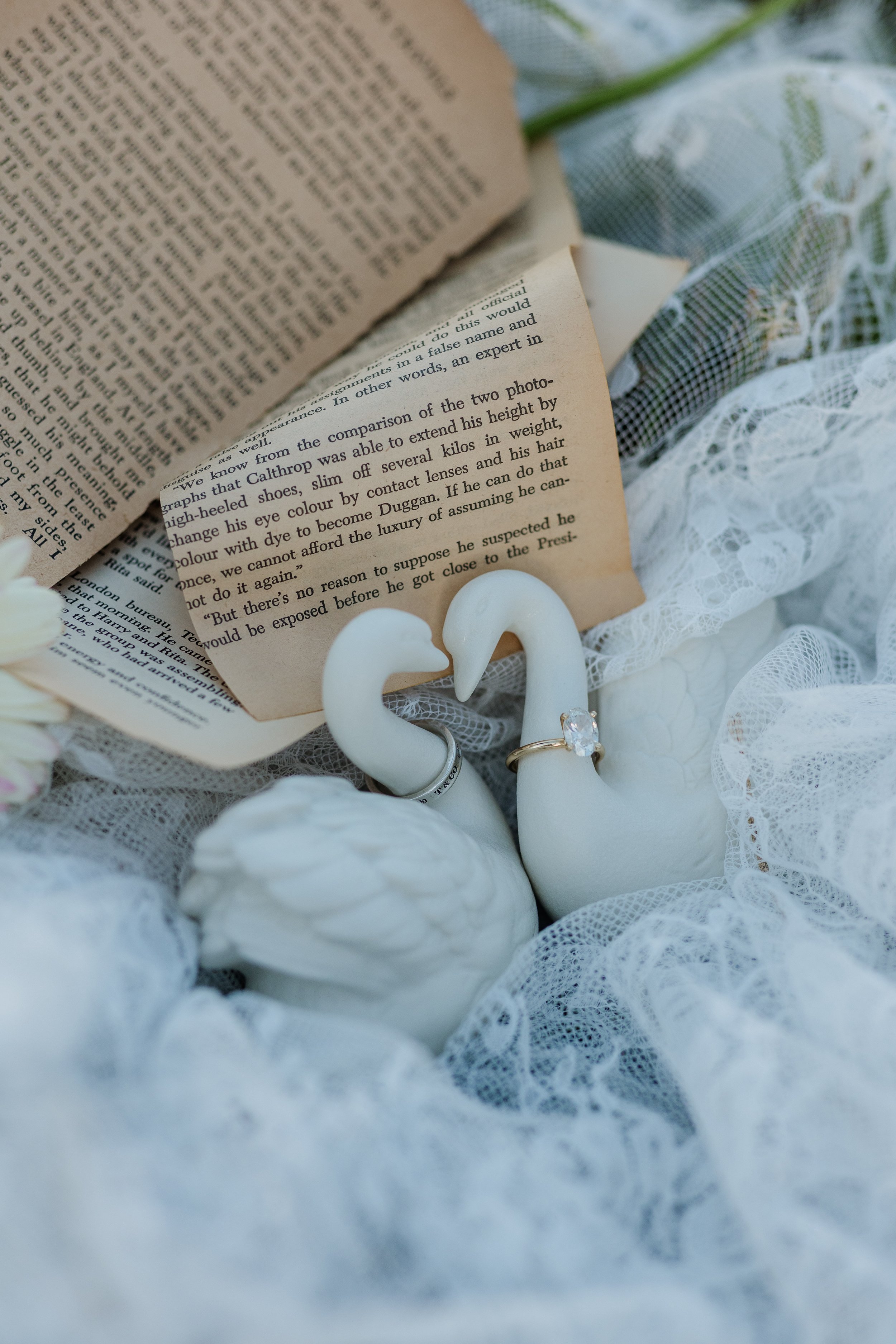Close-up of open pages from a book, a pair of white swan-shaped candles, lace fabric, and jewelry, including a ring with a large gemstone, surrounding the book.