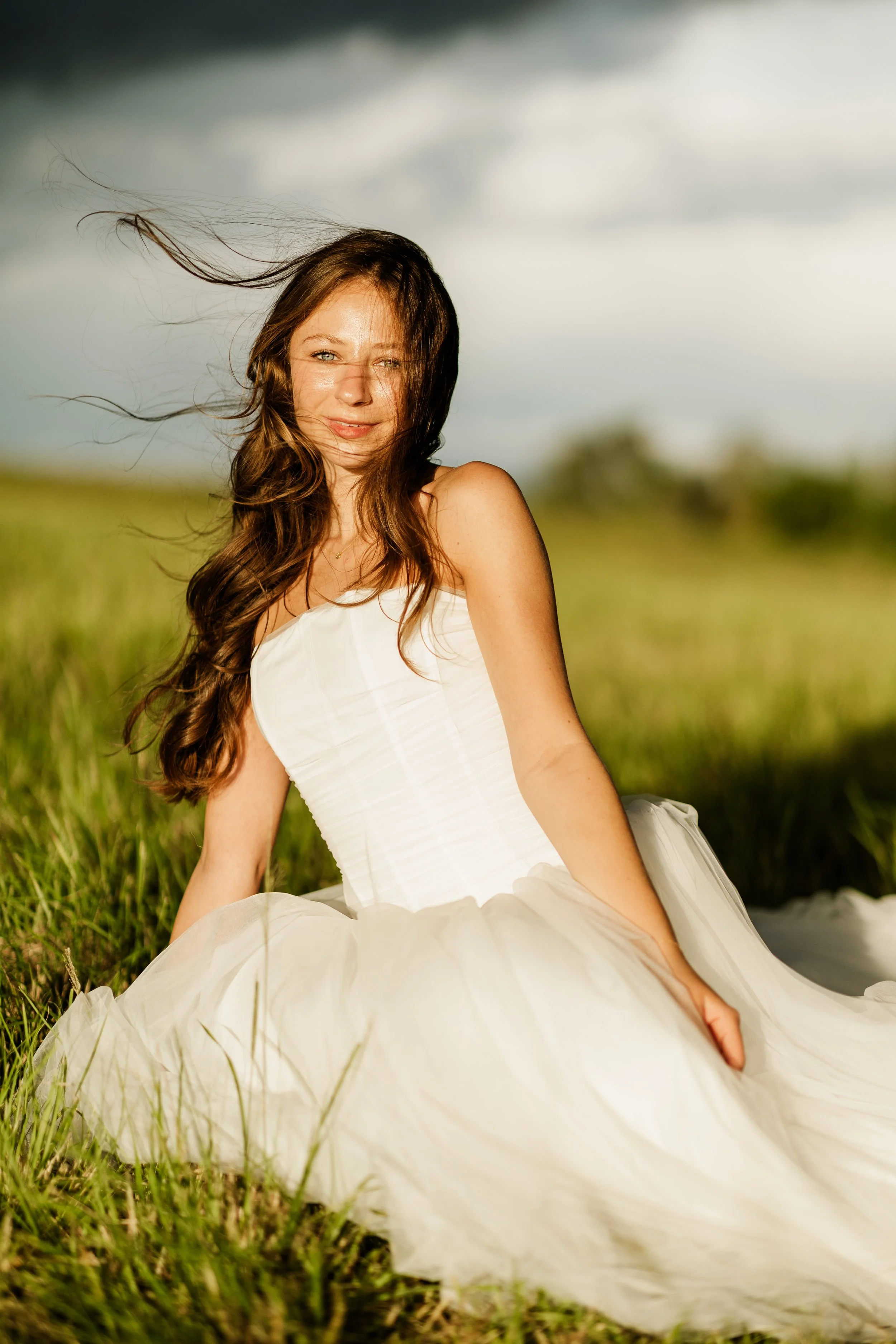 A woman with long brown hair wearing a white dress sitting in a grassy field with a cloudy sky in the background.