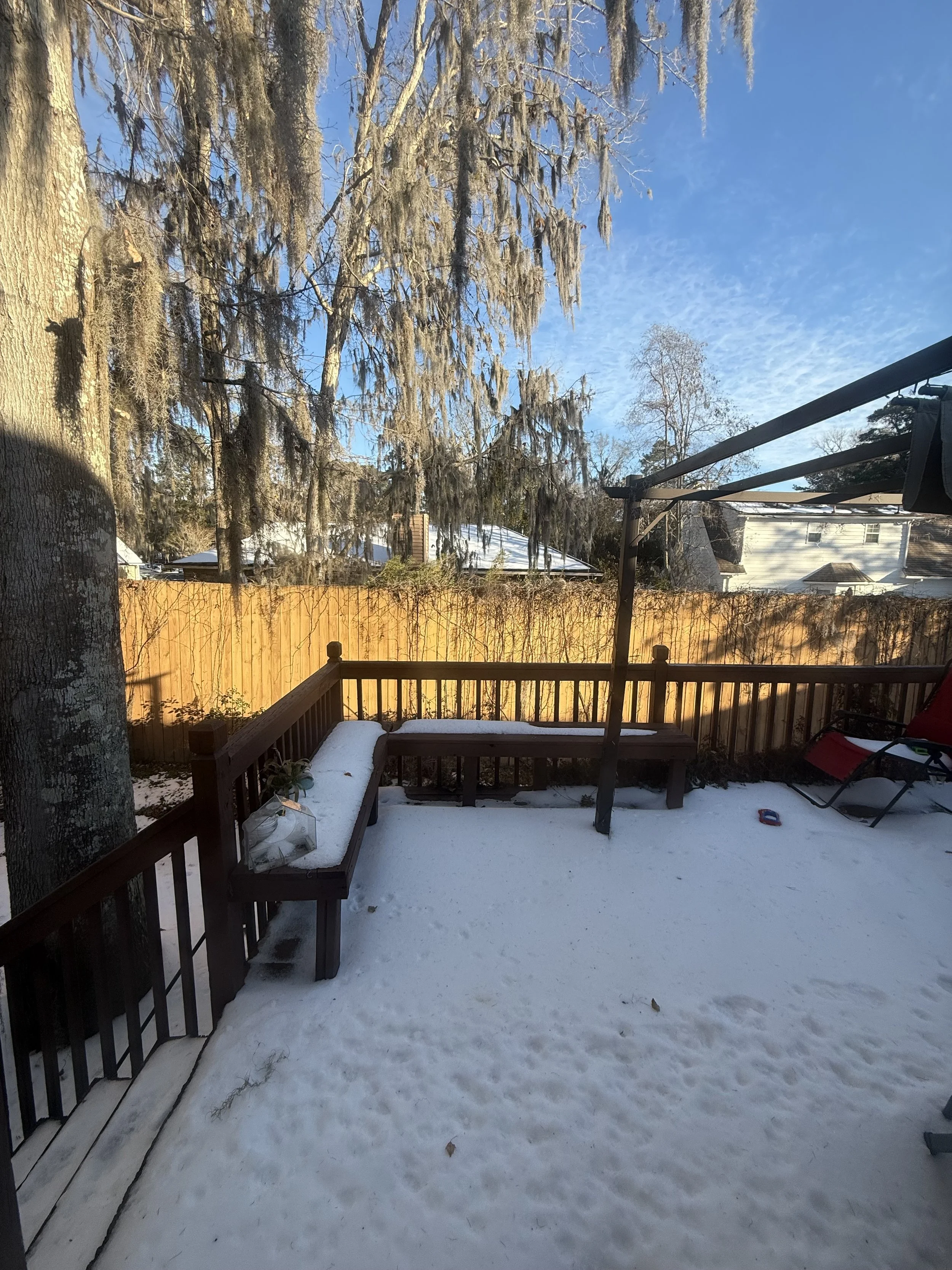 Backyard covered in snow with a wooden deck and bench, large trees with hanging Spanish moss, a wooden fence, and neighboring houses in the distance.