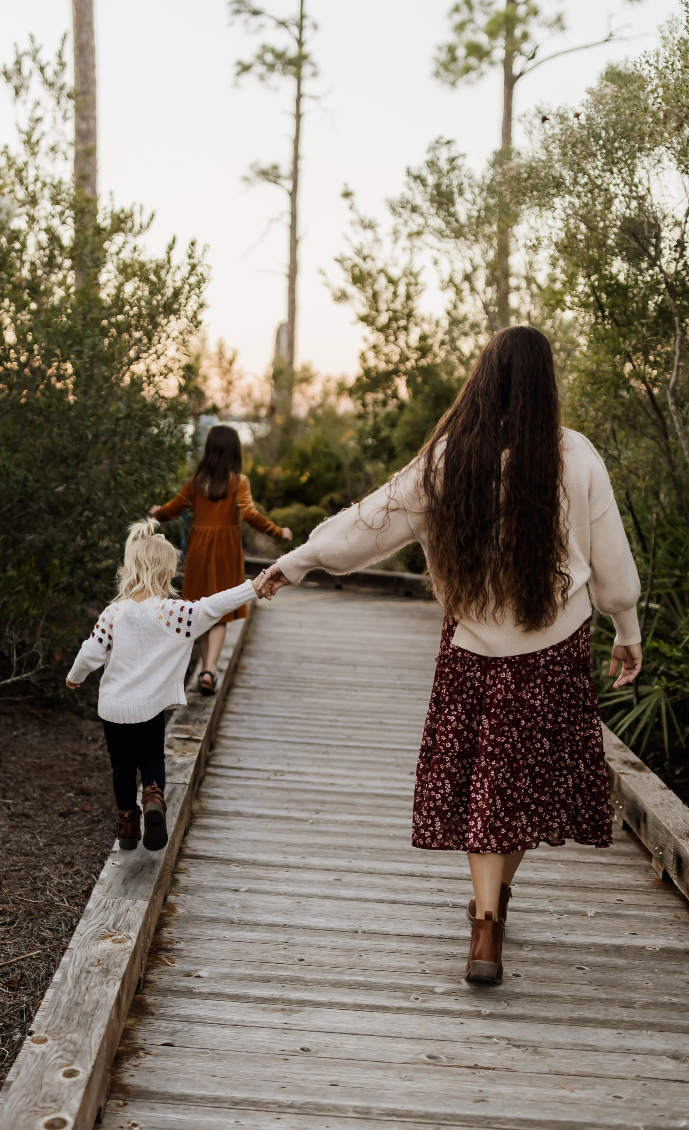 A woman with long, curly brown hair and wearing a white sweater and a floral skirt is holding hands with a young girl in a white sweater and black pants along a wooden pathway surrounded by greenery, with two other girls ahead on the trail near trees