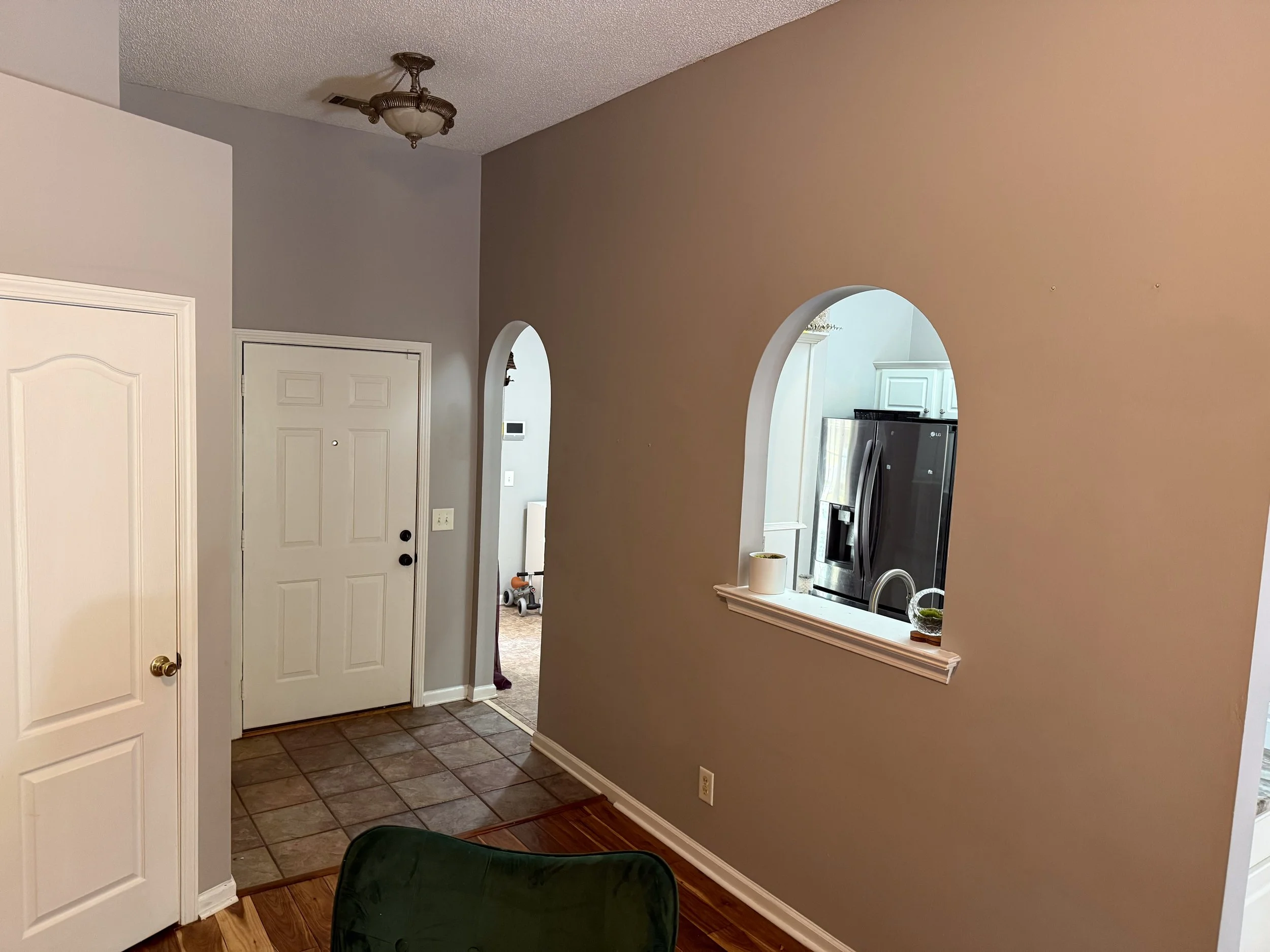 Interior view of a home entryway with a white door and a tiled floor, adjacent to an archway opening to a kitchen. Part of a green chair and a small white table with items on it are visible.
