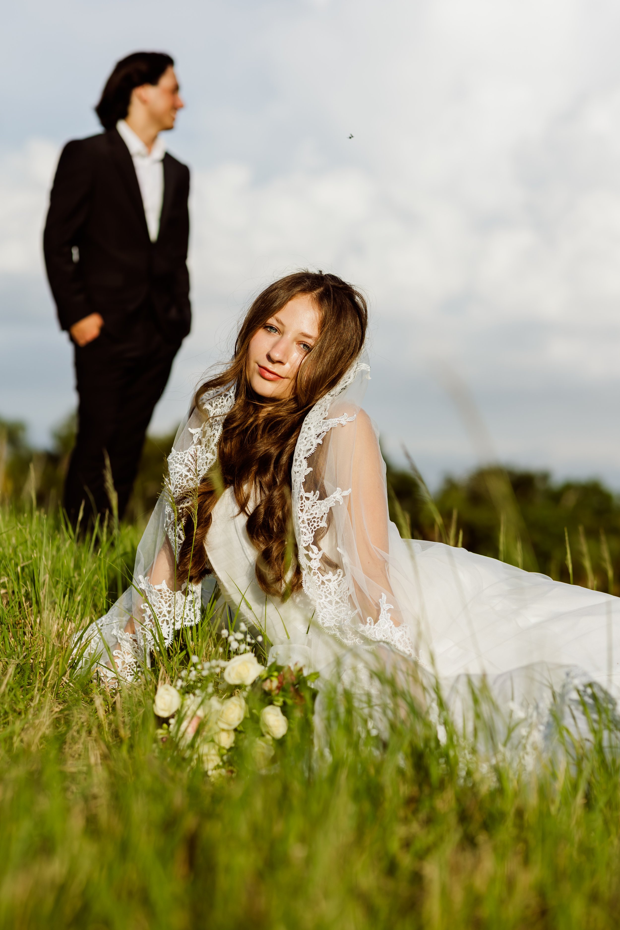 A woman in a wedding dress sitting in a grassy field with white roses, and a man in a black suit standing behind her in the distance, under a cloudy sky.