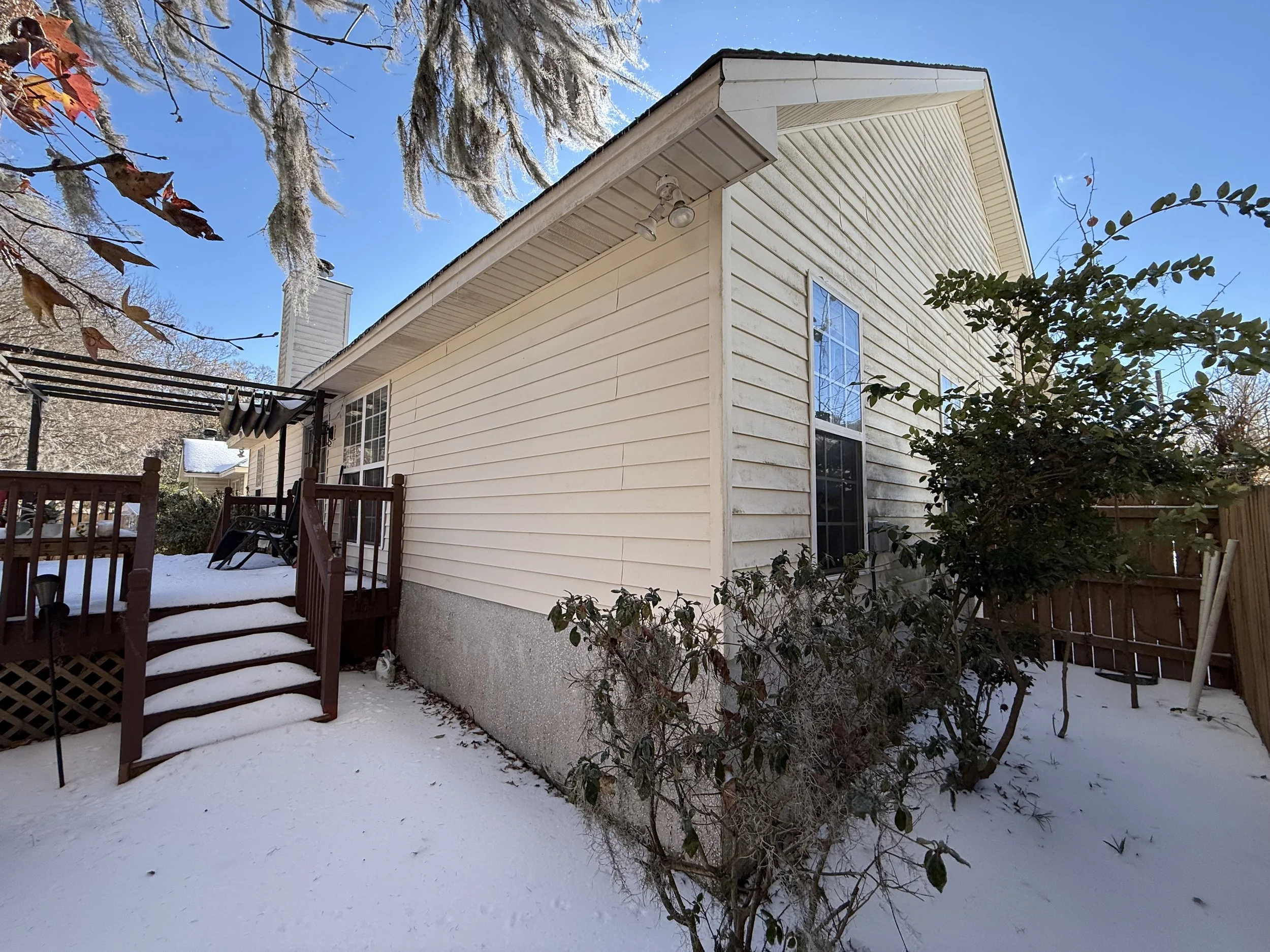Backyard scene with snow-covered ground, a wooden staircase leading to a deck, some bushes, and a house with beige siding and a window. Bare trees and a blue sky are visible in the background.