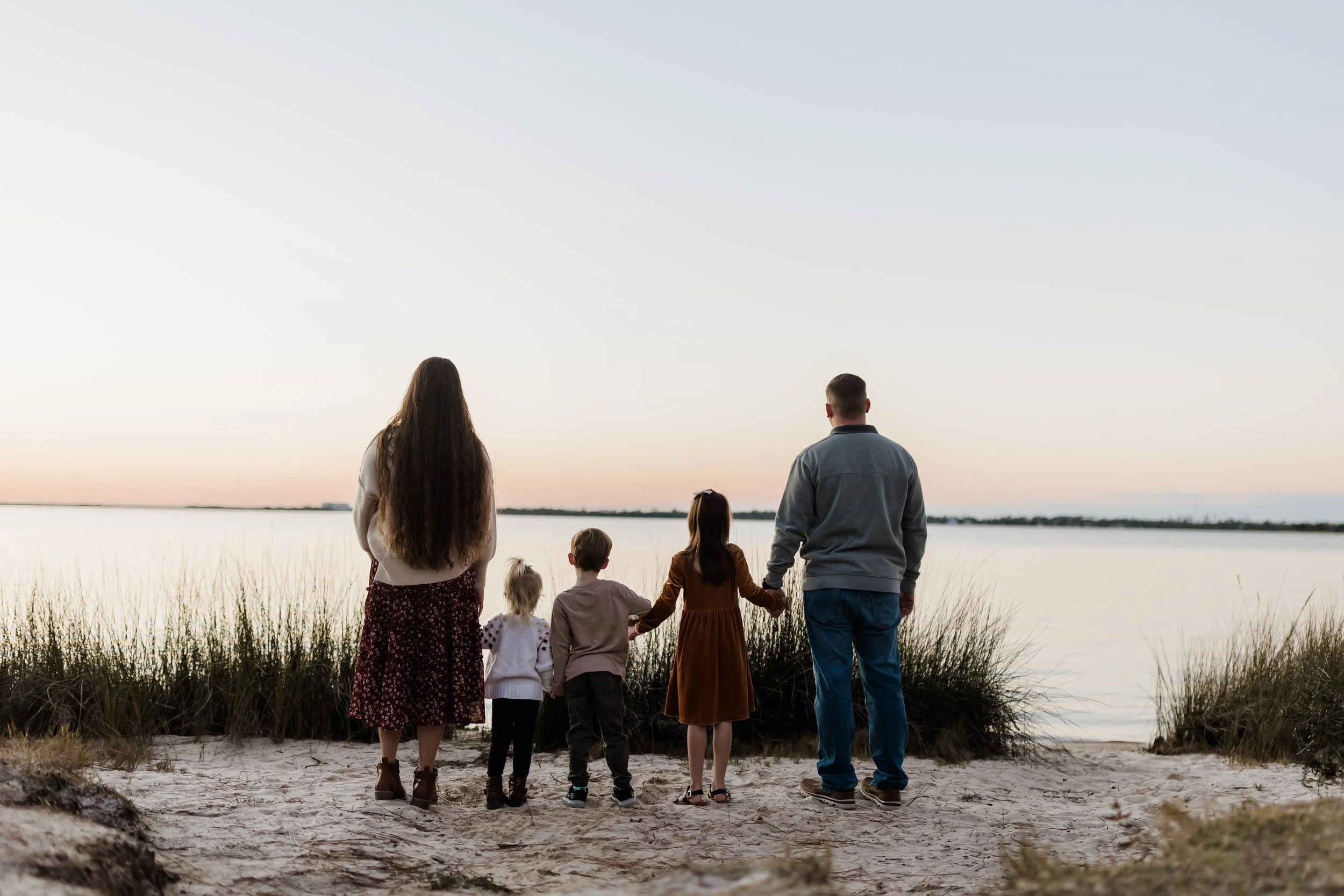 A group of seven people, including adults and children, standing on a sandy beach near a body of water during sunset, looking across the water.