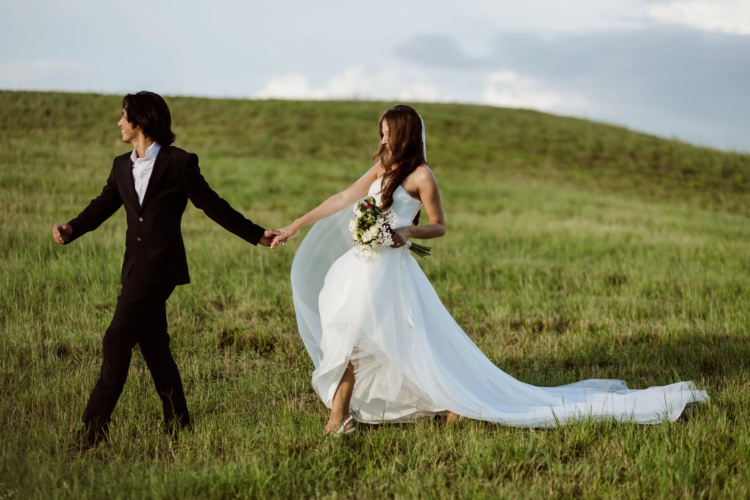 A bride in a white wedding gown holding a bouquet, walking through a grassy field with a groom in a black suit leading her by the hand.