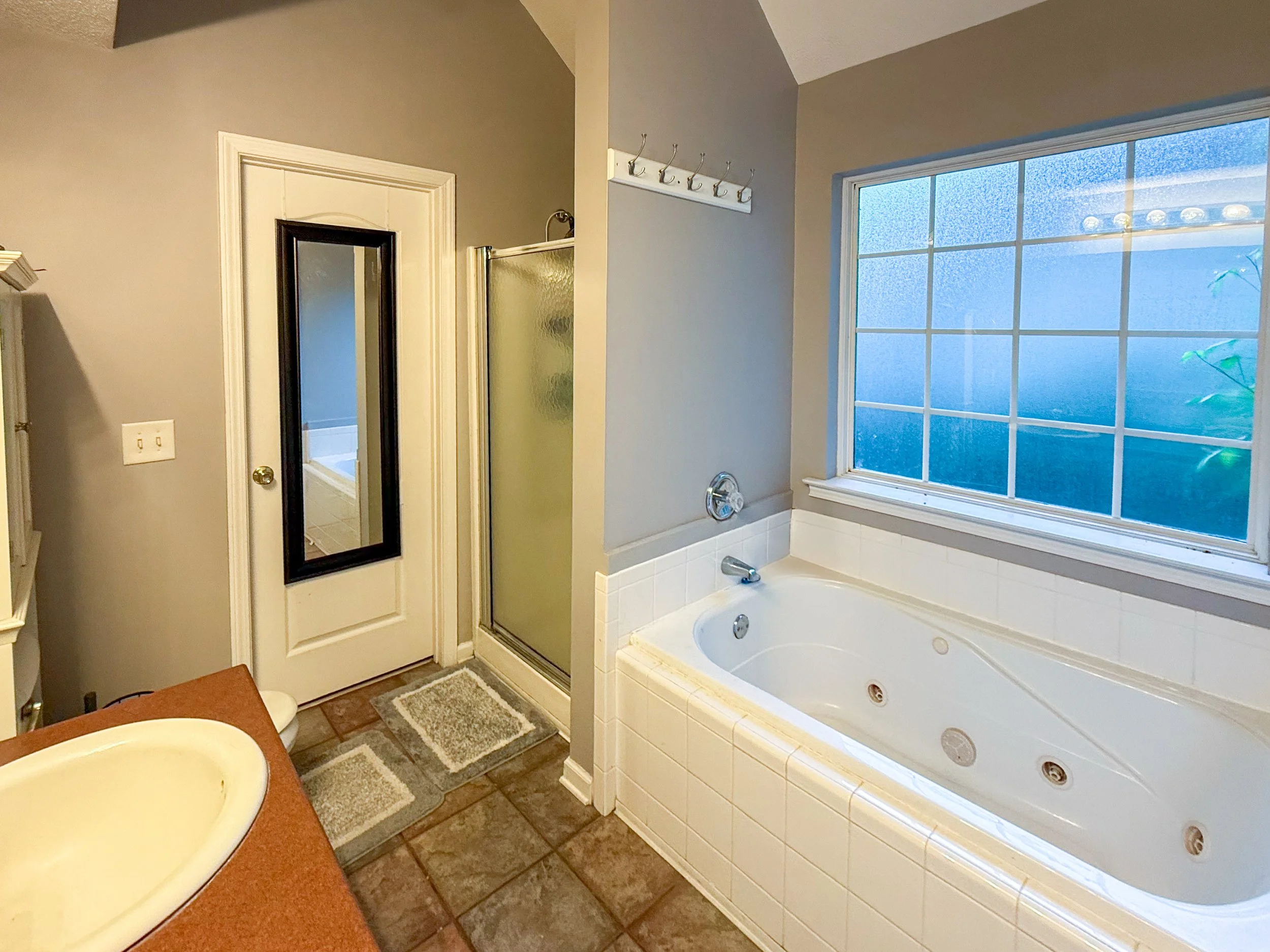 A bathroom with a large window, a jetted bathtub, a shower stall, a mirror, and a small sink with a countertop.