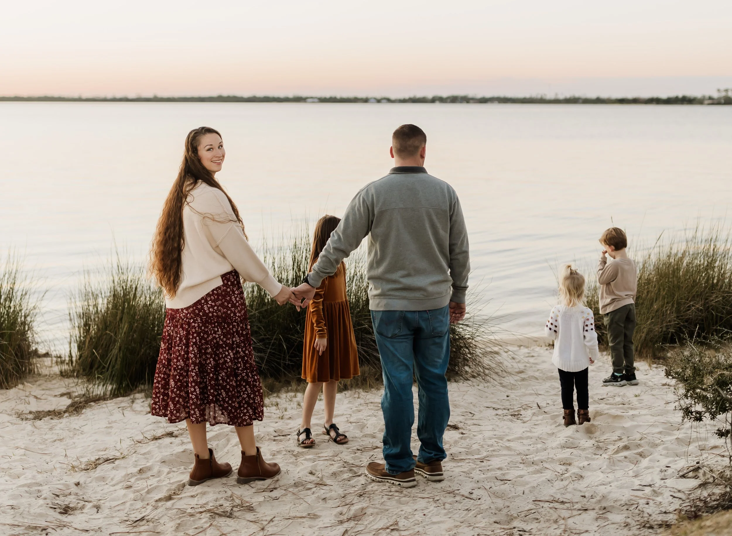 A group of people holding hands and walking on a sandy beach near water, with some looking out at the water and others facing away. The sky is pale, suggesting early morning or late evening.