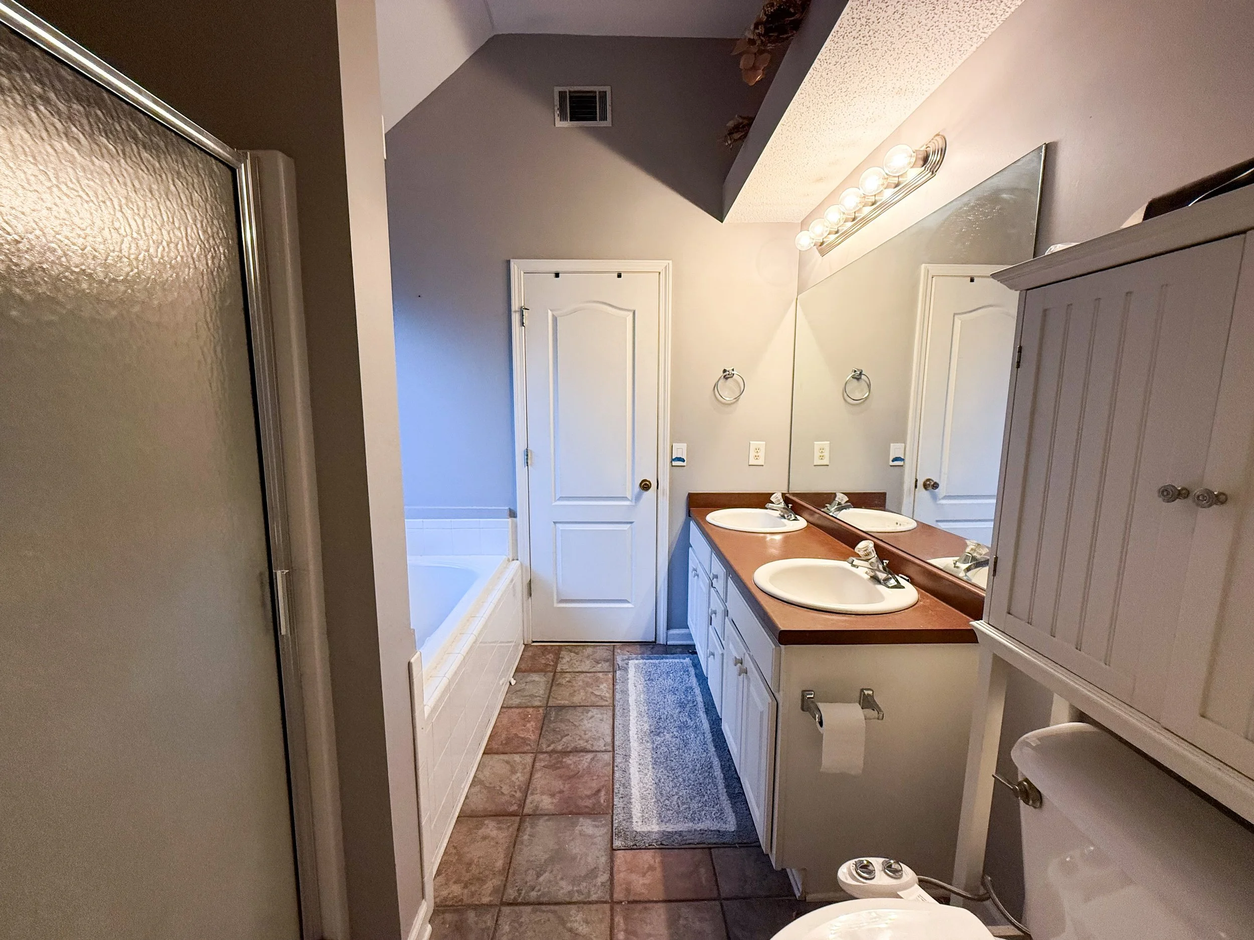 A bathroom with a double sink vanity, large mirror, light fixtures, tile floor, toilet, cabinet, and shower, with a door at the back.