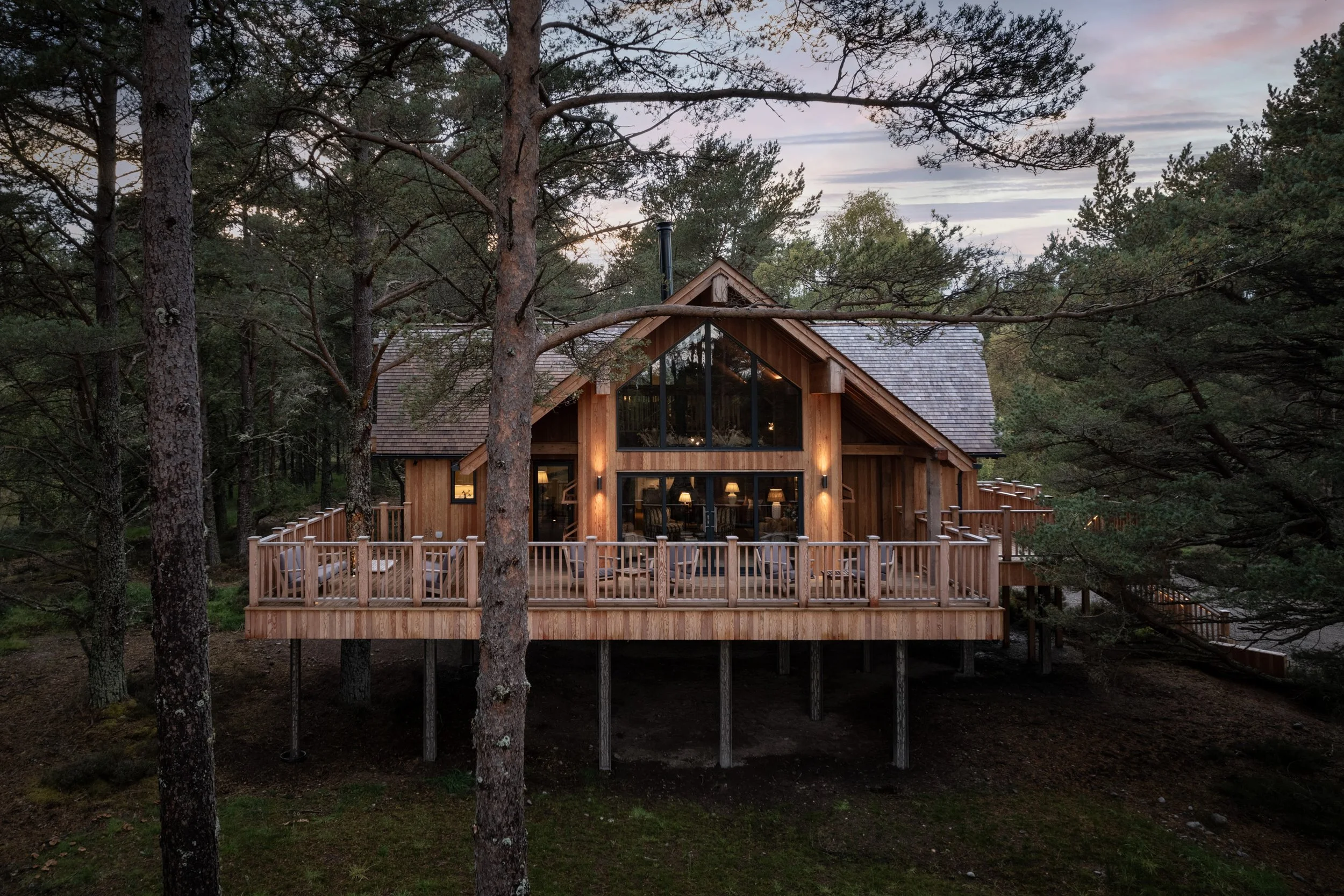 A wooden house elevated on stilts, surrounded by trees, with a large deck and large windows looking out onto the forested area.