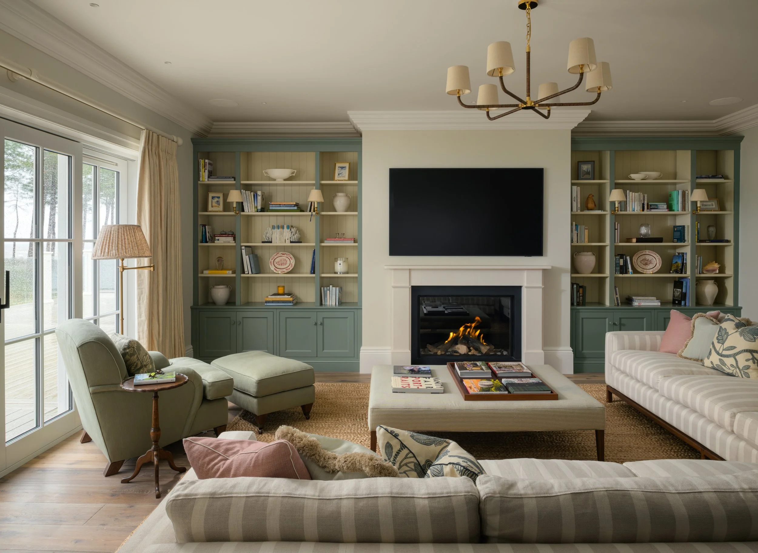Living room with a fireplace, a large flat-screen TV, beige couches with patterned pillows, a footrest and small tables, bookshelves on either side of the fireplace, a sliding glass door with curtains, and hardwood floors.