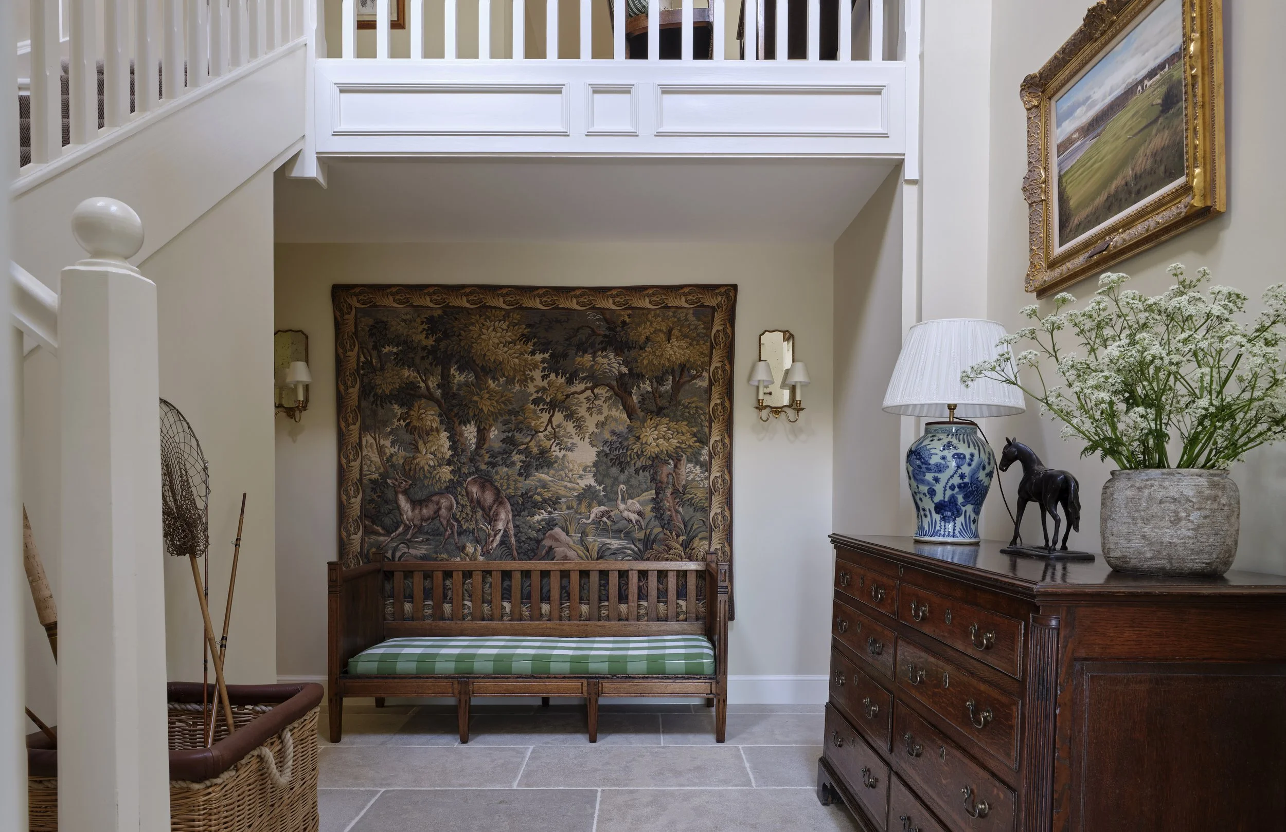 Comfortable entryway with vintage decor, featuring a large tapestry of a forest scene with animals on the back wall, a wooden bench with green striped cushion, and an antique wooden dresser topped with a blue and white lamp, a black horse figurine, and a potted plant with white flowers.