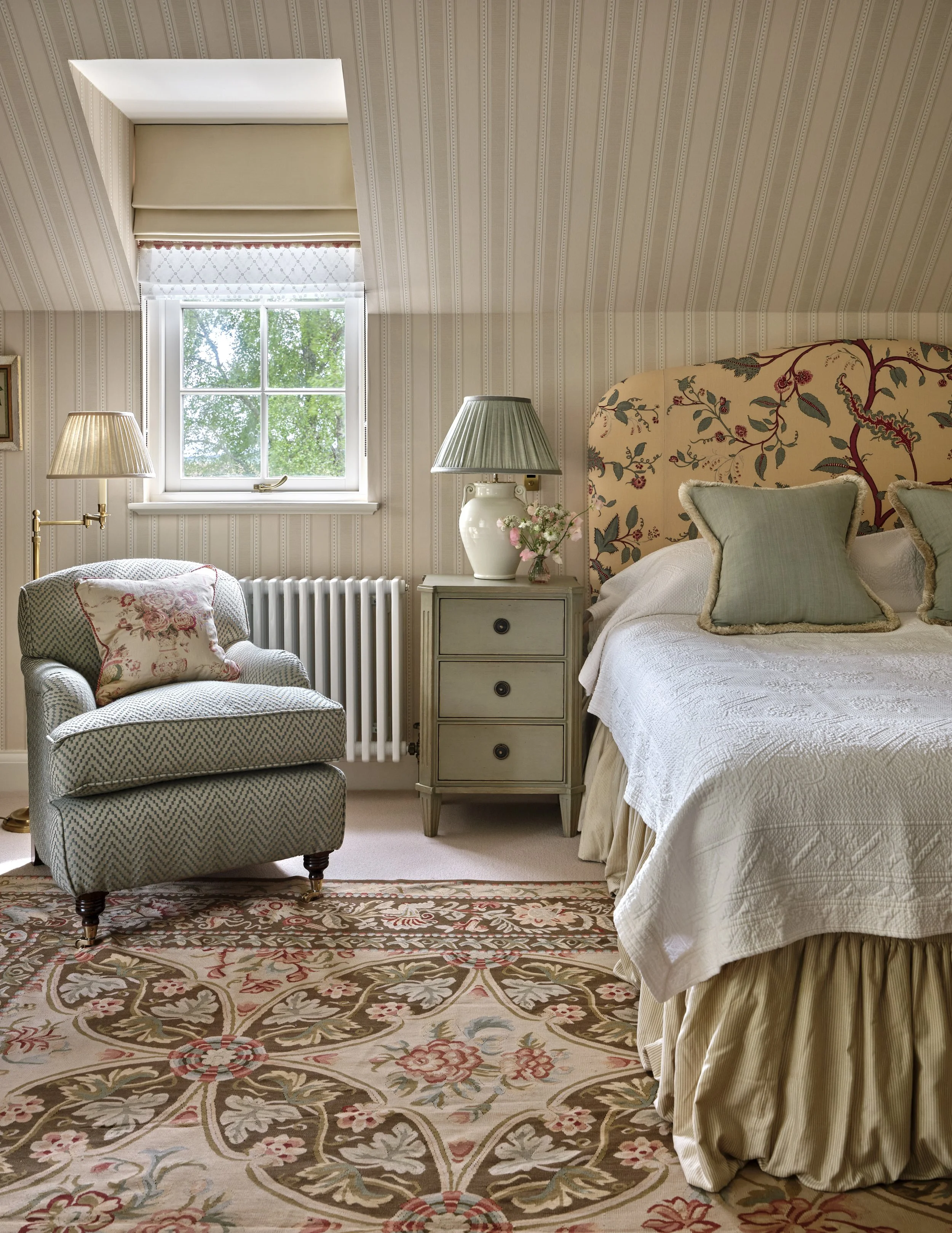 A cozy bedroom with a window, armchair with floral pillow, small side table with a lamp and flowers, and a bed with a floral headboard and decorative pillows, all in soft pastel tones.