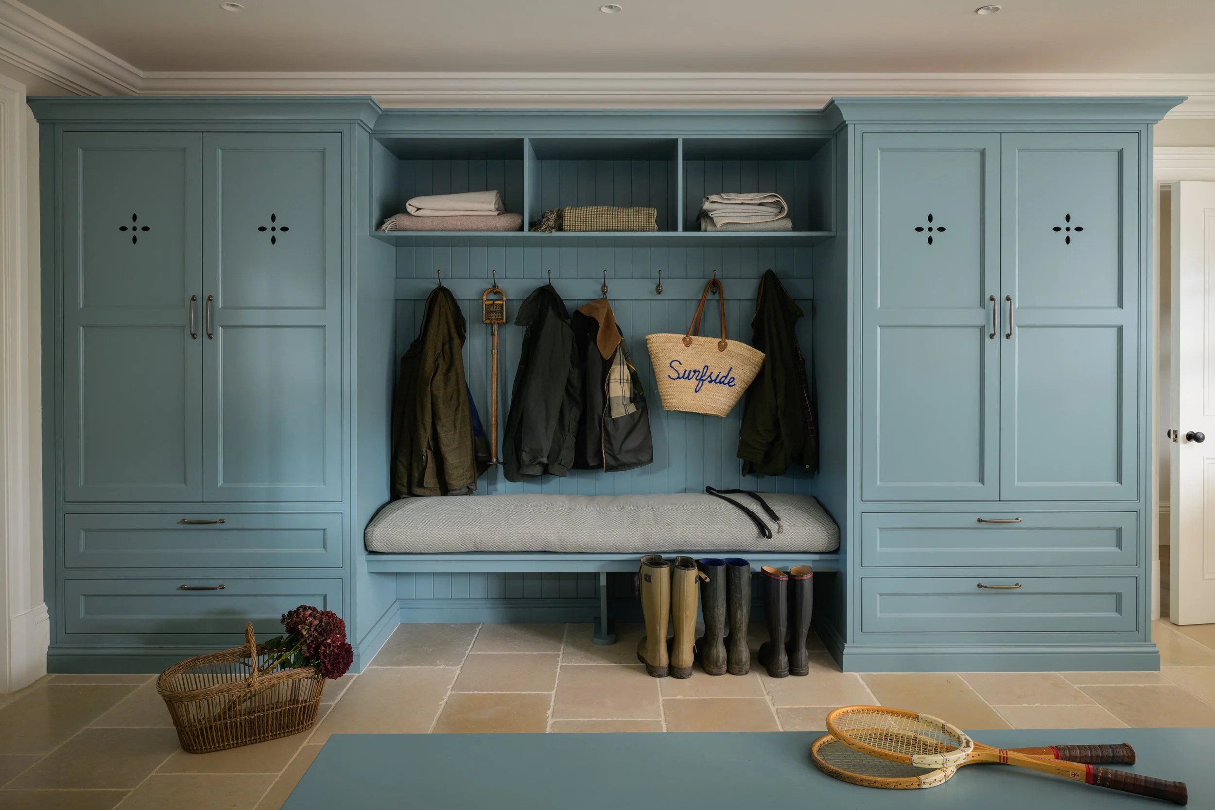 A blue built-in hall tree with shelves, hooks, and cabinets, holding coats, bags, and folded towels. There are rubber boots and a seat with a cushion underneath. A basket with flowers is on the beige tiled floor, and tennis rackets are on a blue table in the foreground.