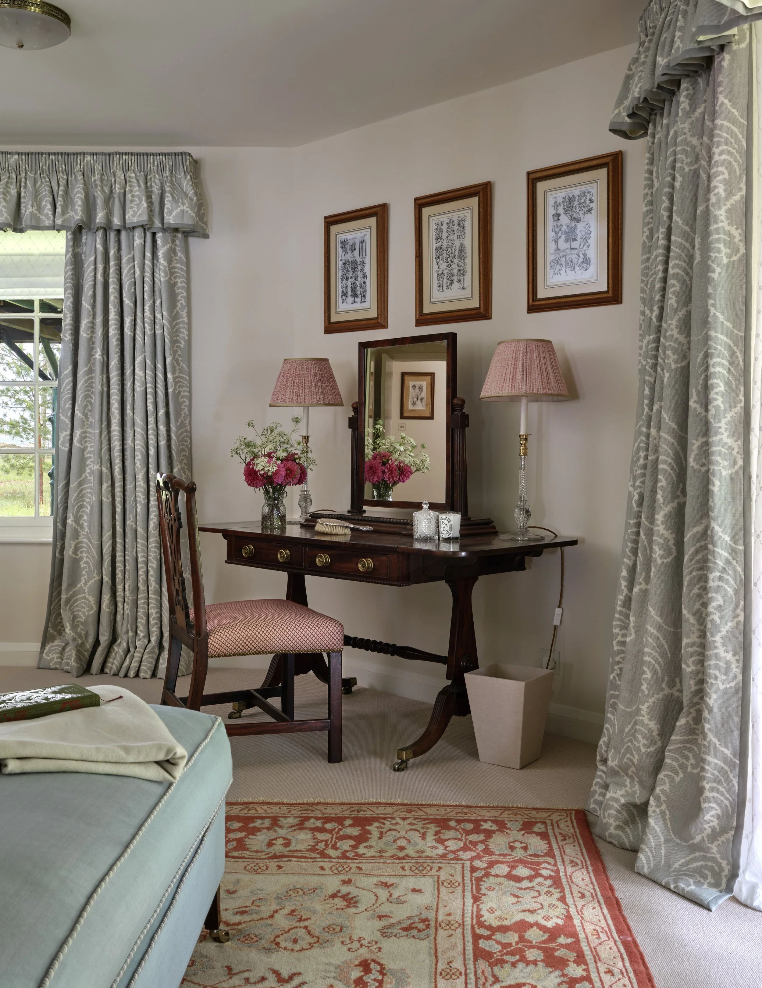 A bedroom corner with a wooden vanity table, mirror, and two pink lamps. There are framed botanical prints above and floral arrangements on the table. A window with patterned drapes is to the left, and an upholstered chair and part of a bed are visib